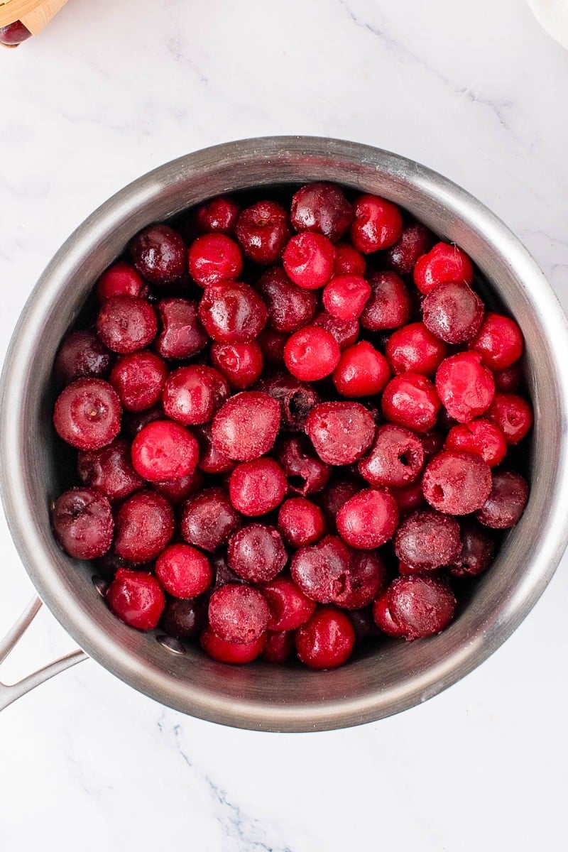 A stainless steel pot filled with frozen, pitted cherries sits on a white marble surface.