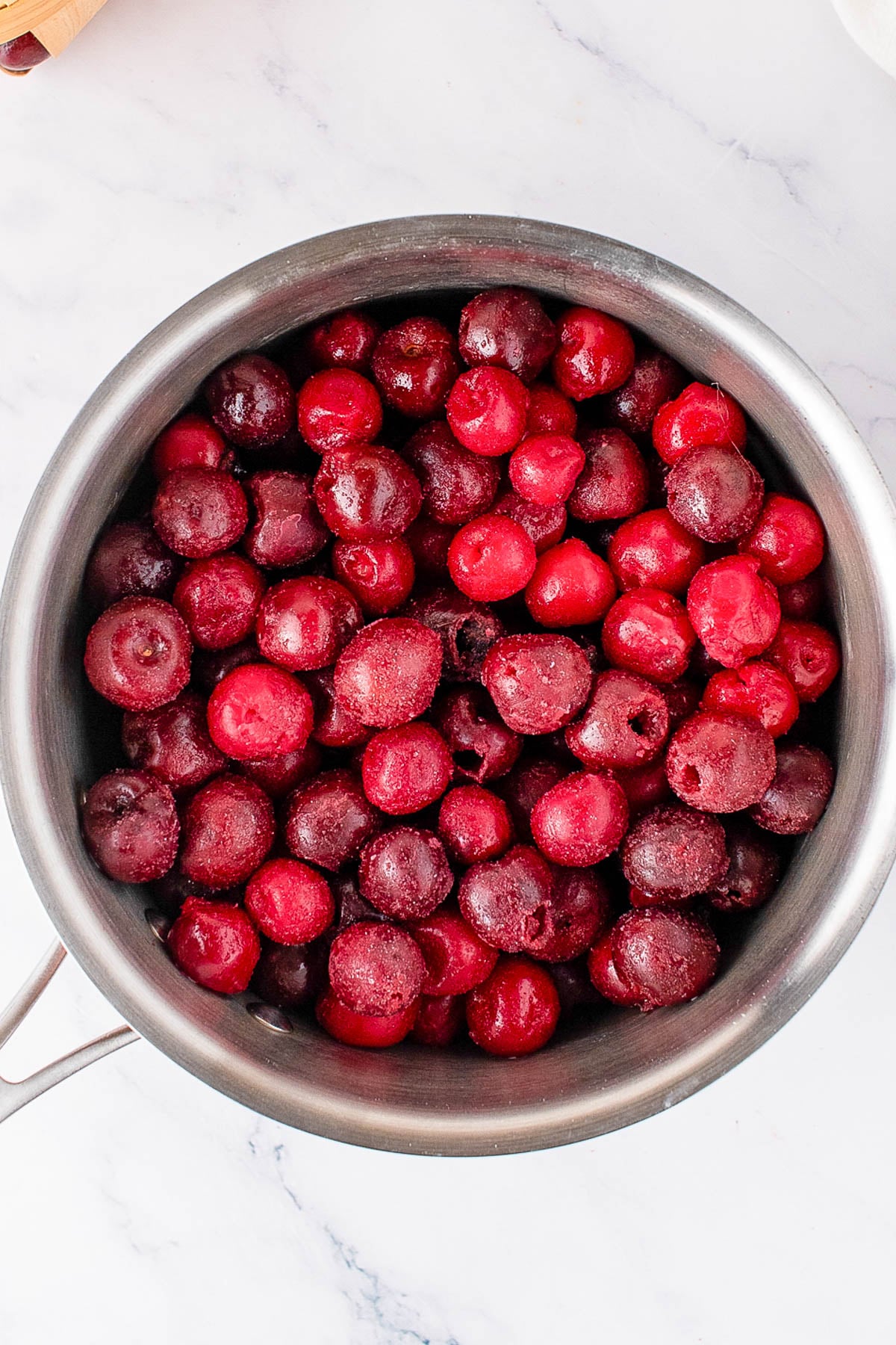 A stainless steel pot filled with frozen, pitted cherries sits on a white marble surface.