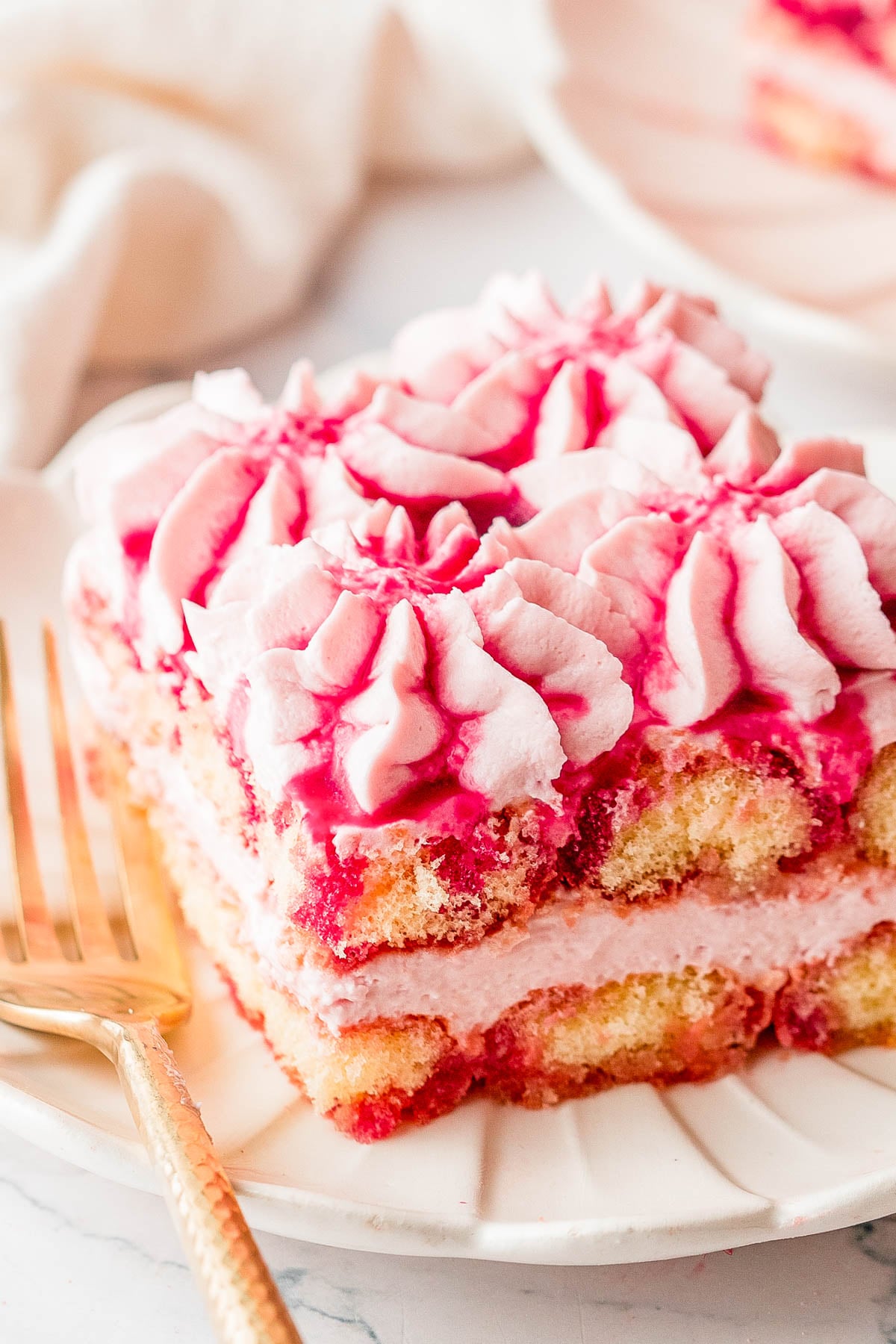 A slice of layered cake with pink and white frosting, served on a white plate with a gold fork.