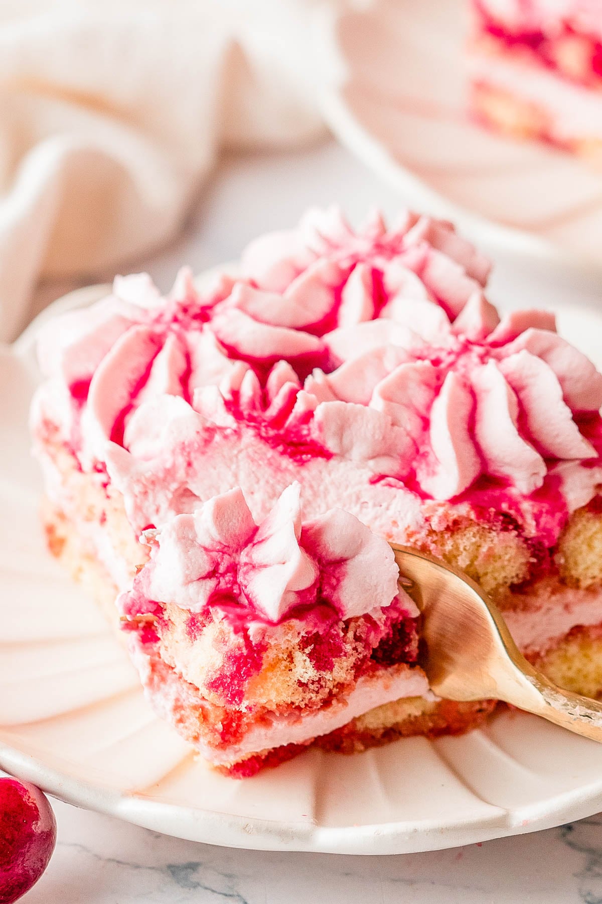 A fork cuts into a slice of layered dessert with pink whipped topping and raspberry sauce, served on a white plate.