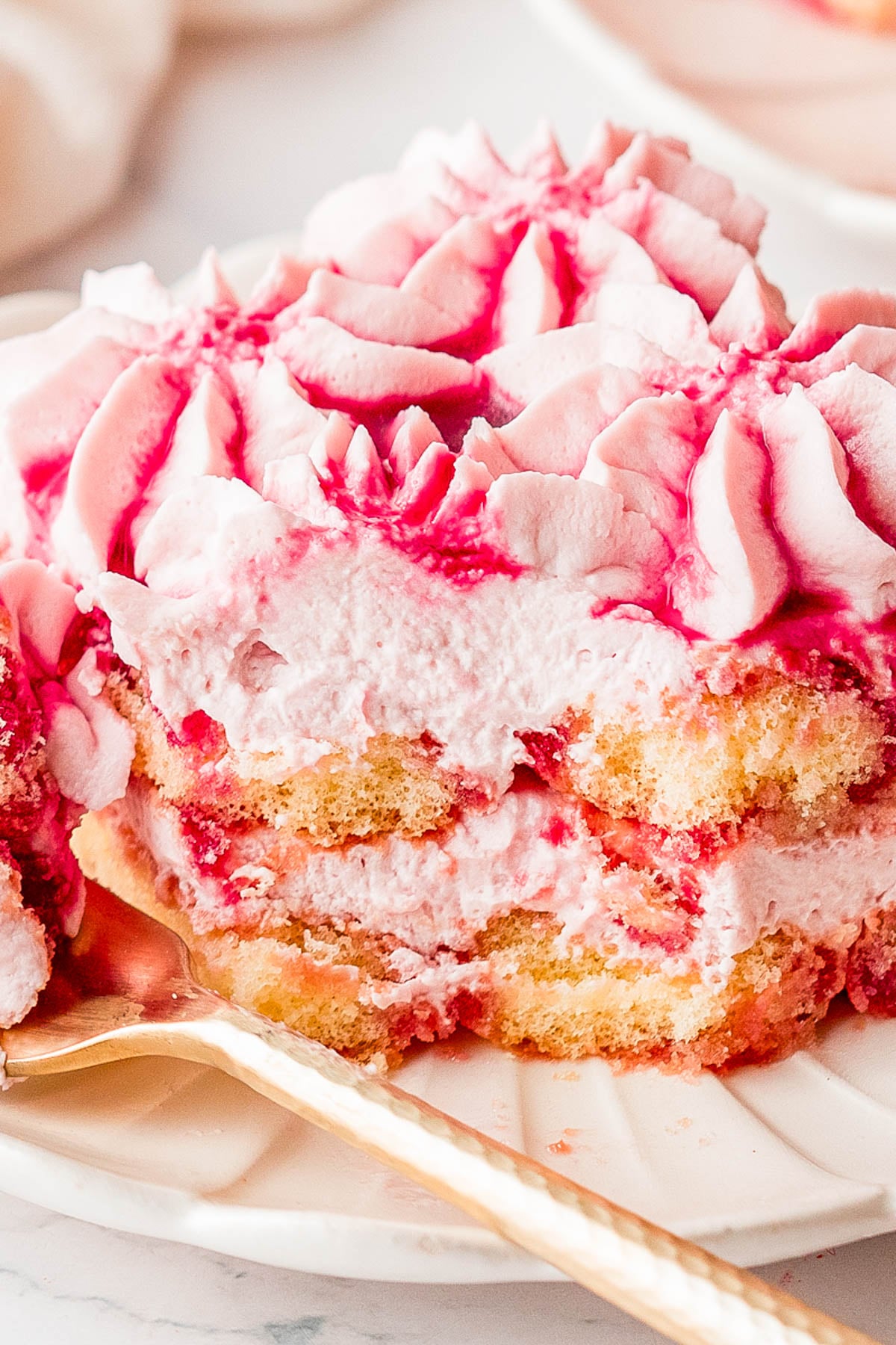 A close-up of a slice of pink raspberry tiramisu with layers of cream and ladyfinger cookies, served on a white plate with a gold spoon.