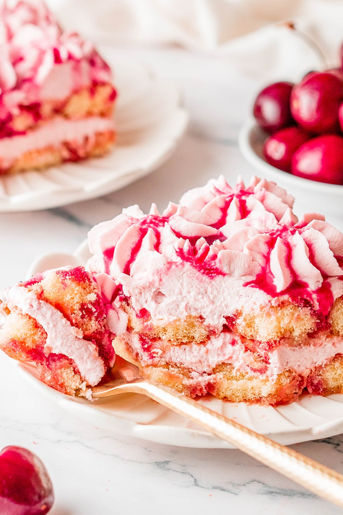 A slice of cherry tiramisu with pink cream and cherry sauce is served on a plate with a fork; fresh cherries are visible in the background.