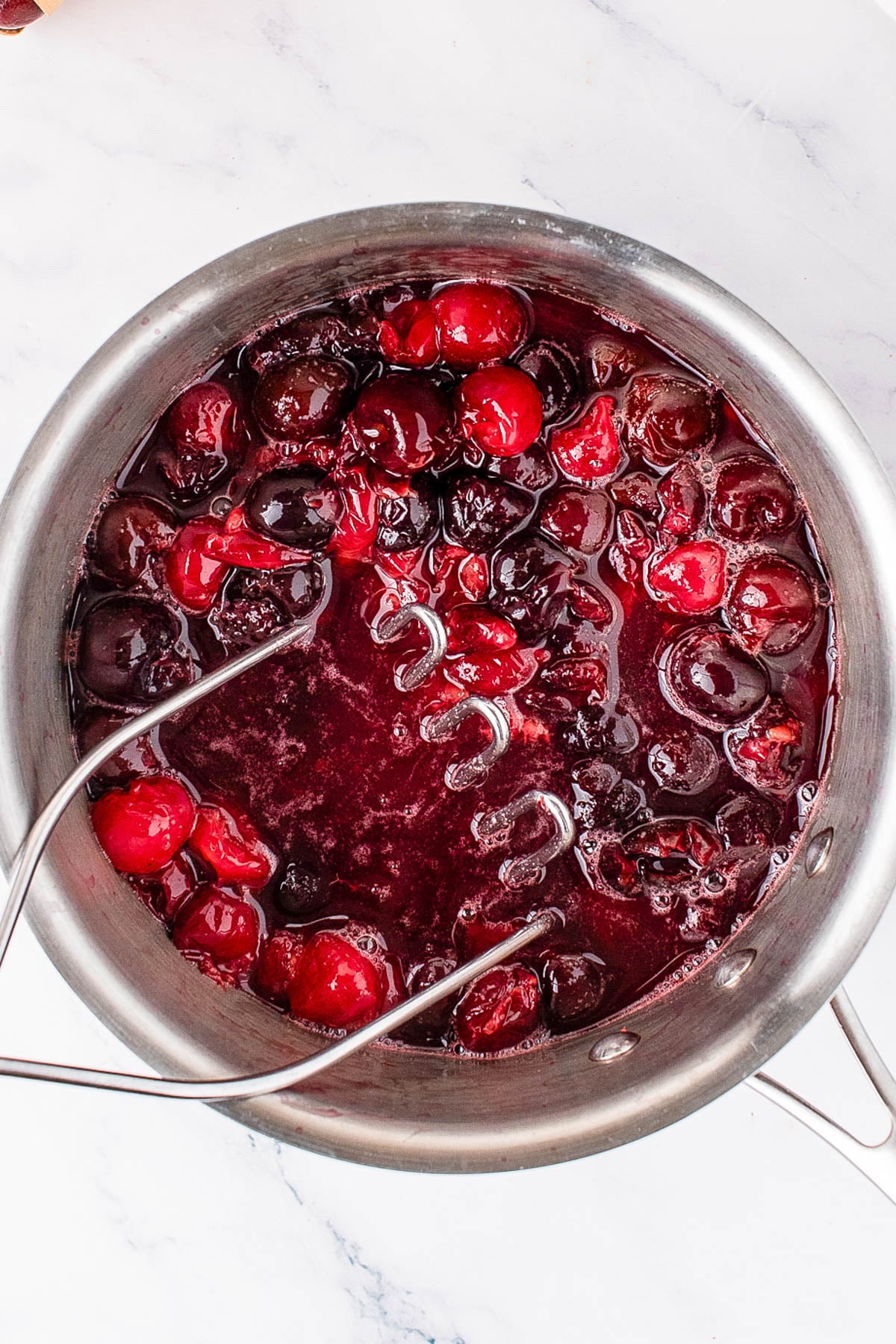 A stainless steel pot filled with whole cherries and cherry sauce, with a potato masher partially submerged in the mixture on a white marble surface.