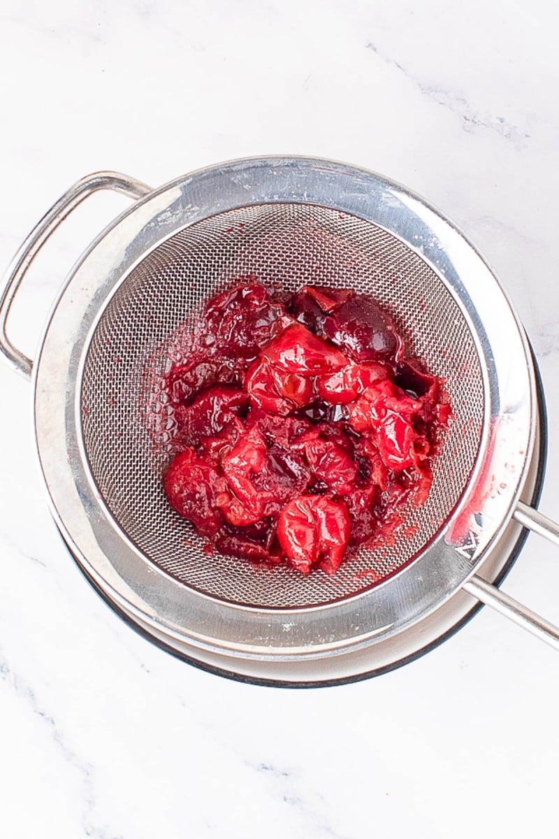 A metal strainer holds cooked, mashed plums with juice draining into a bowl below, on a white marble surface.
