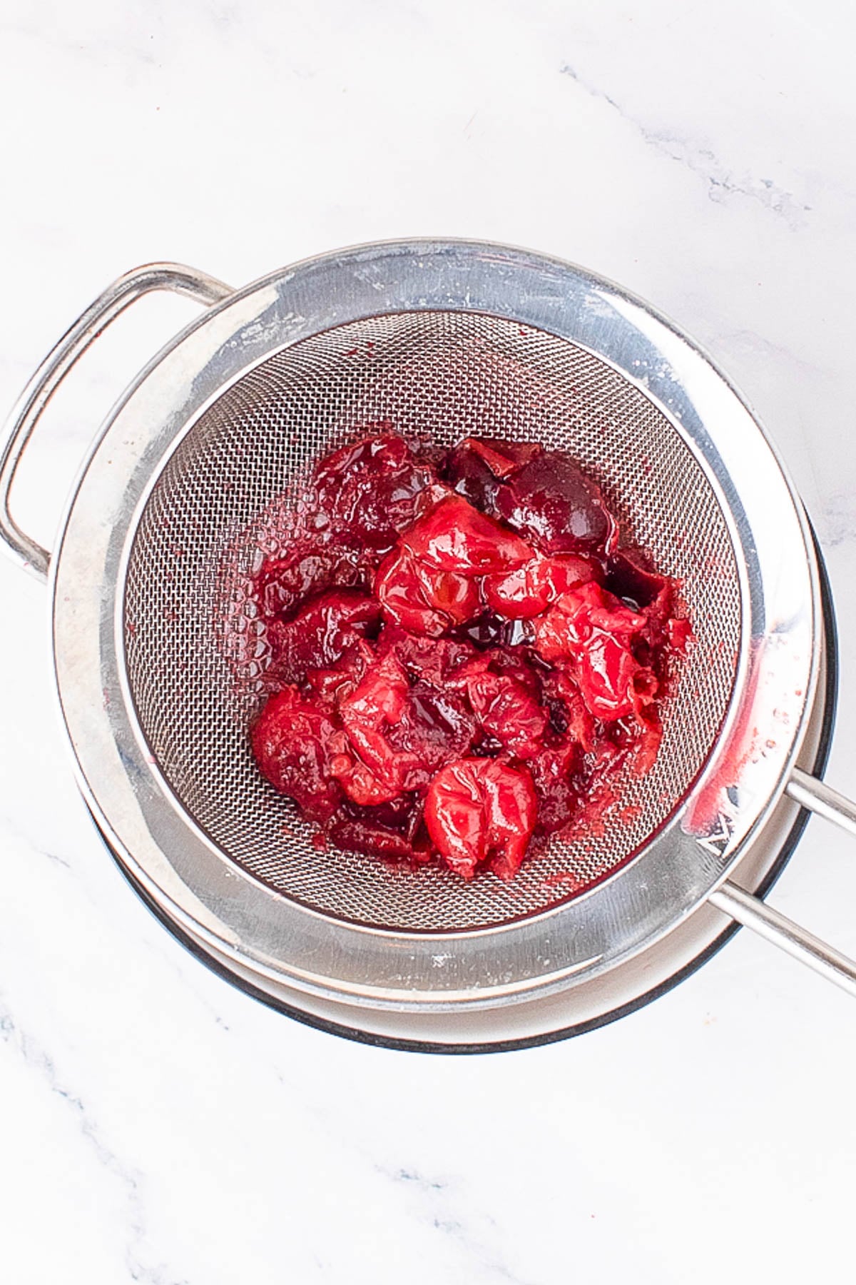 A metal strainer holds cooked, mashed plums with juice draining into a bowl below, on a white marble surface.