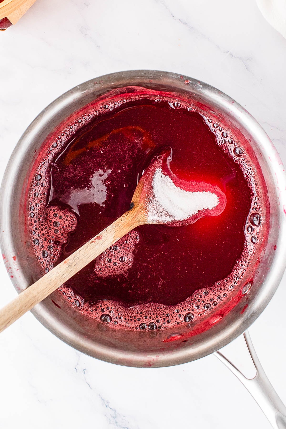 A saucepan filled with red liquid being stirred with a wooden spoon, viewed from above on a white surface.