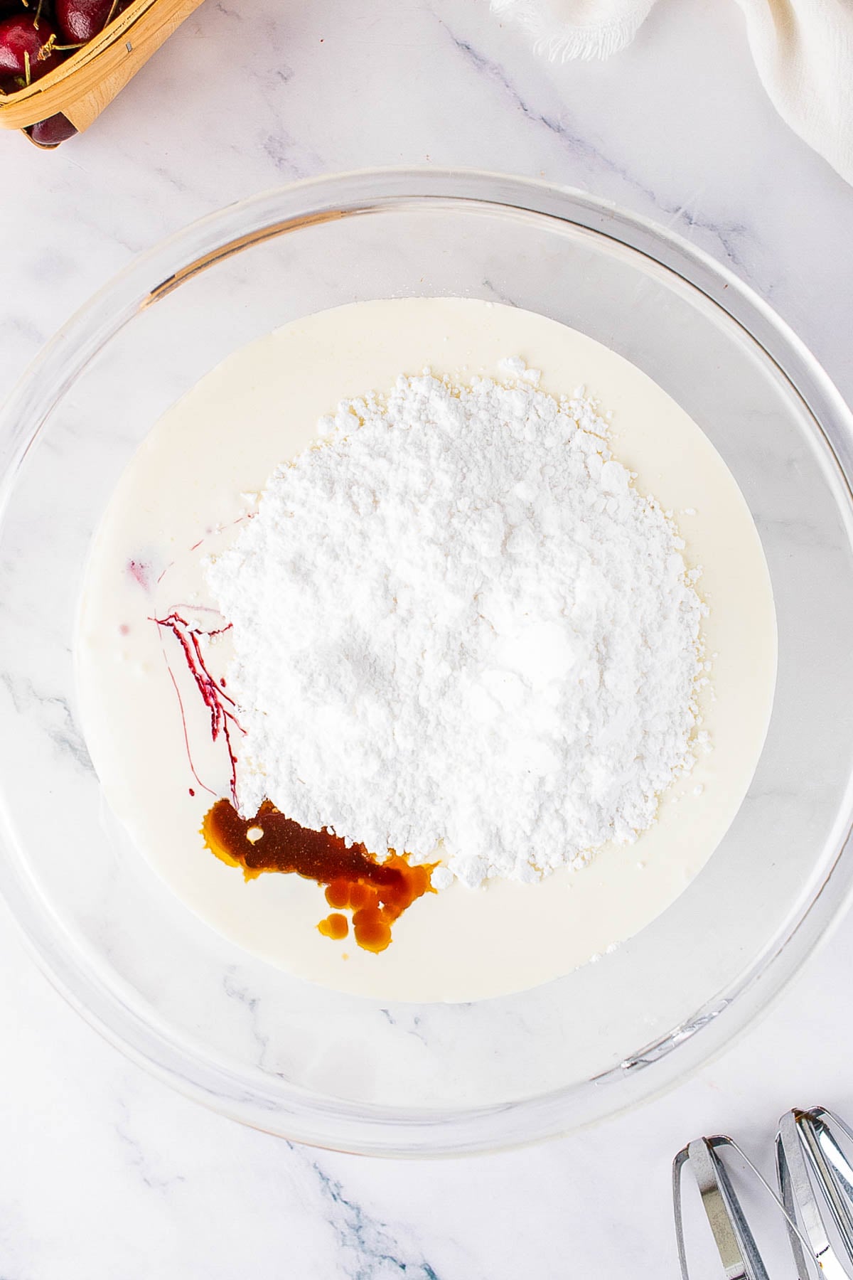 A glass bowl contains heavy cream, powdered sugar, vanilla extract, and a small amount of red food coloring, ready for mixing.