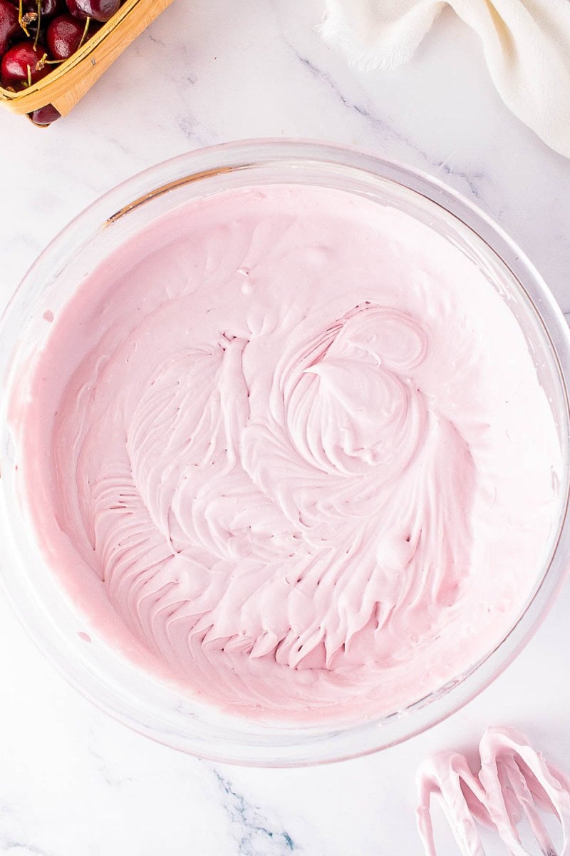A glass bowl filled with pink whipped mixture, with electric mixer beaters coated in the same mixture resting nearby on a marble surface.