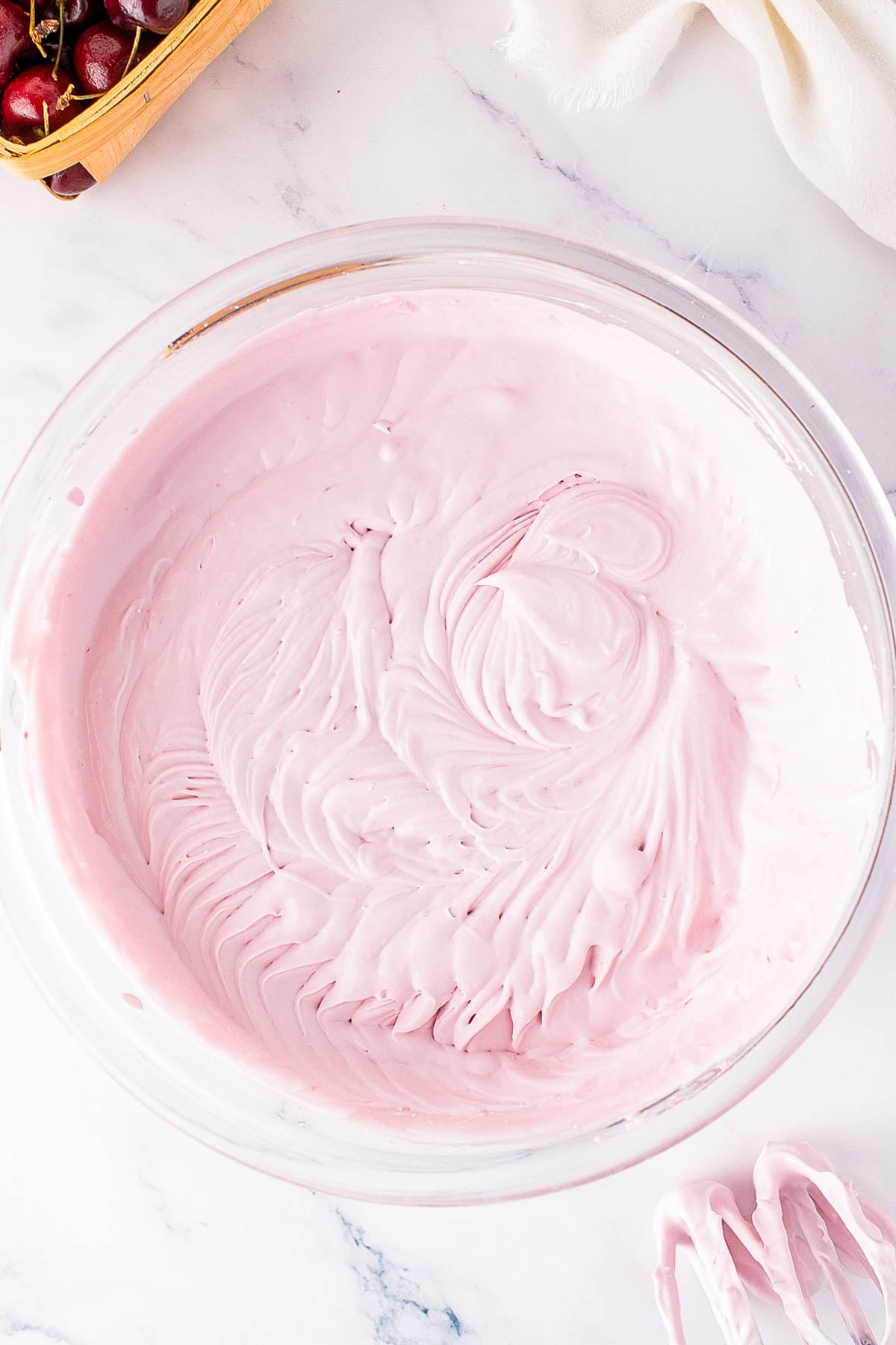 A glass bowl filled with pink whipped mixture, with electric mixer beaters coated in the same mixture resting nearby on a marble surface.