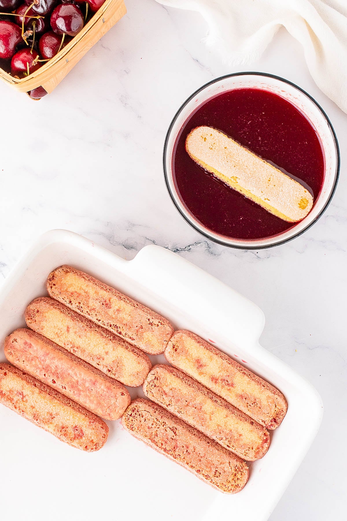 Ladyfingers being dipped in a bowl of cherry liquid, with a baking dish containing soaked ladyfingers and a basket of cherries nearby on a marble surface.