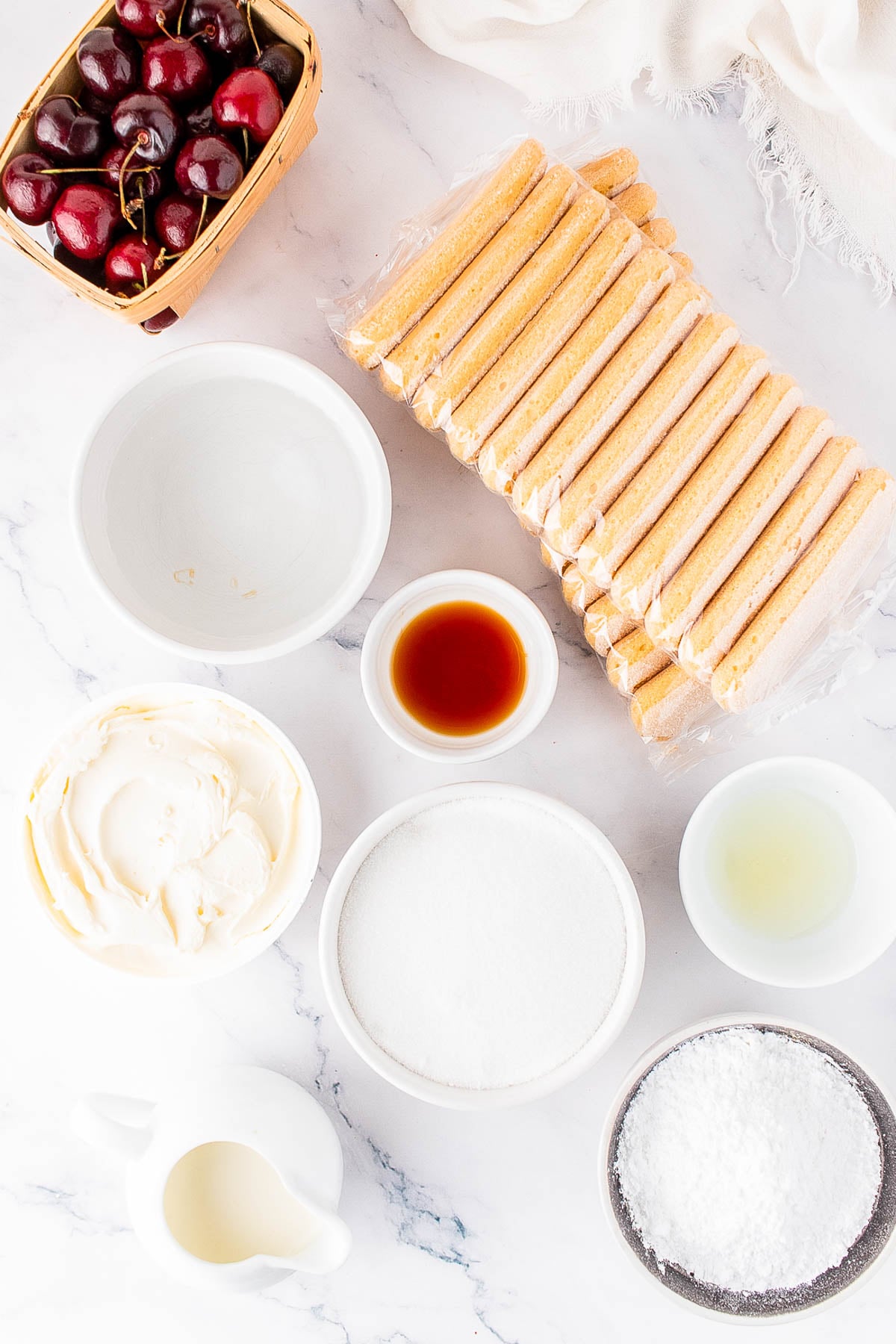 Assorted tiramisu ingredients on a white surface, including cherries, ladyfingers, mascarpone, sugar, vanilla, lemon juice, powdered sugar, and cream in bowls.