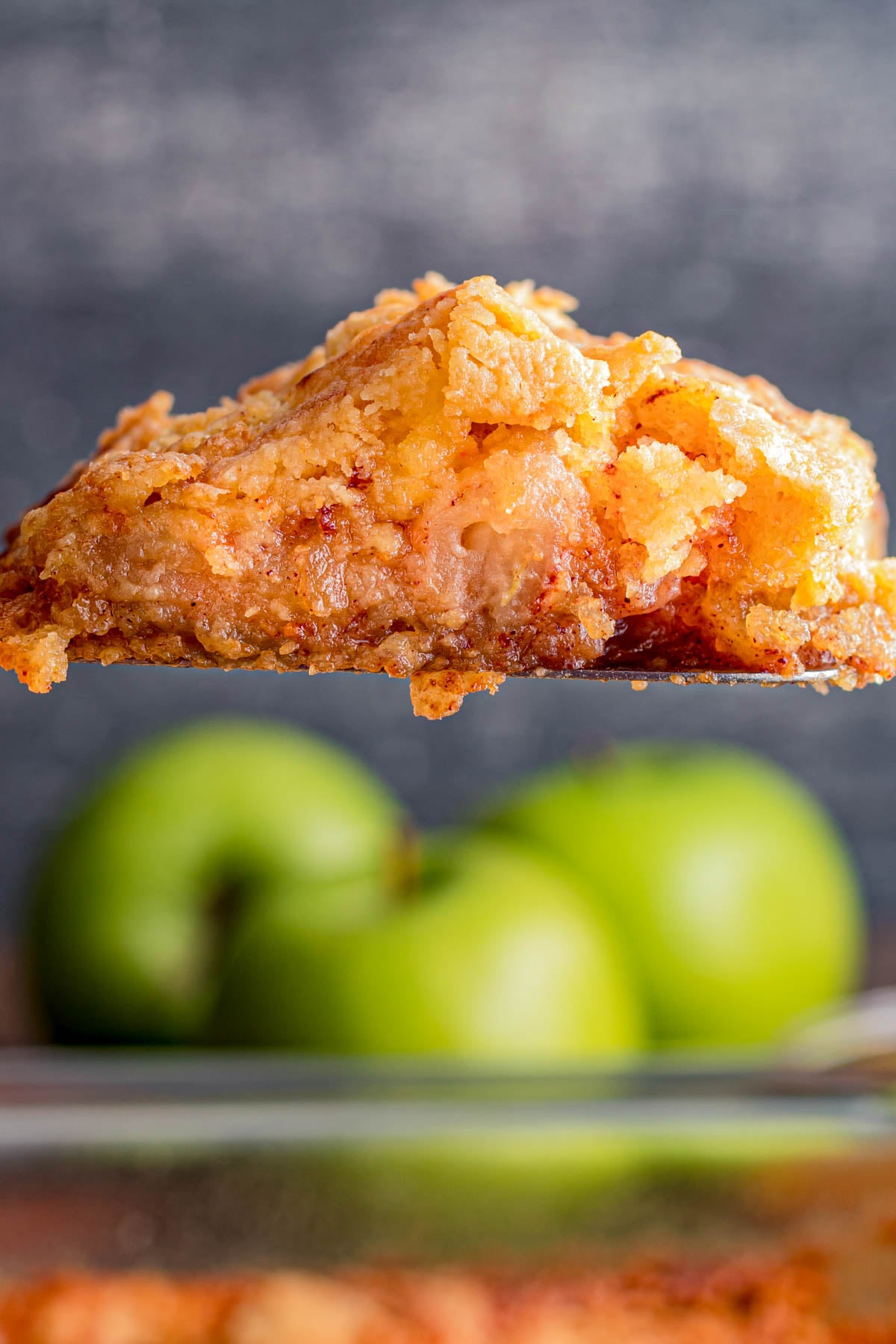 A close-up of a serving of apple cobbler on a spatula, with green apples blurred in the background.