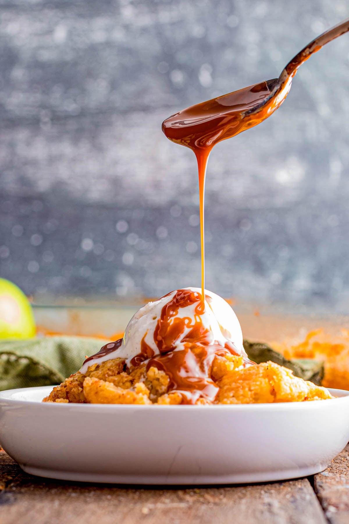 A spoon drizzles caramel sauce over a scoop of vanilla ice cream on top of an apple dessert in a white bowl.