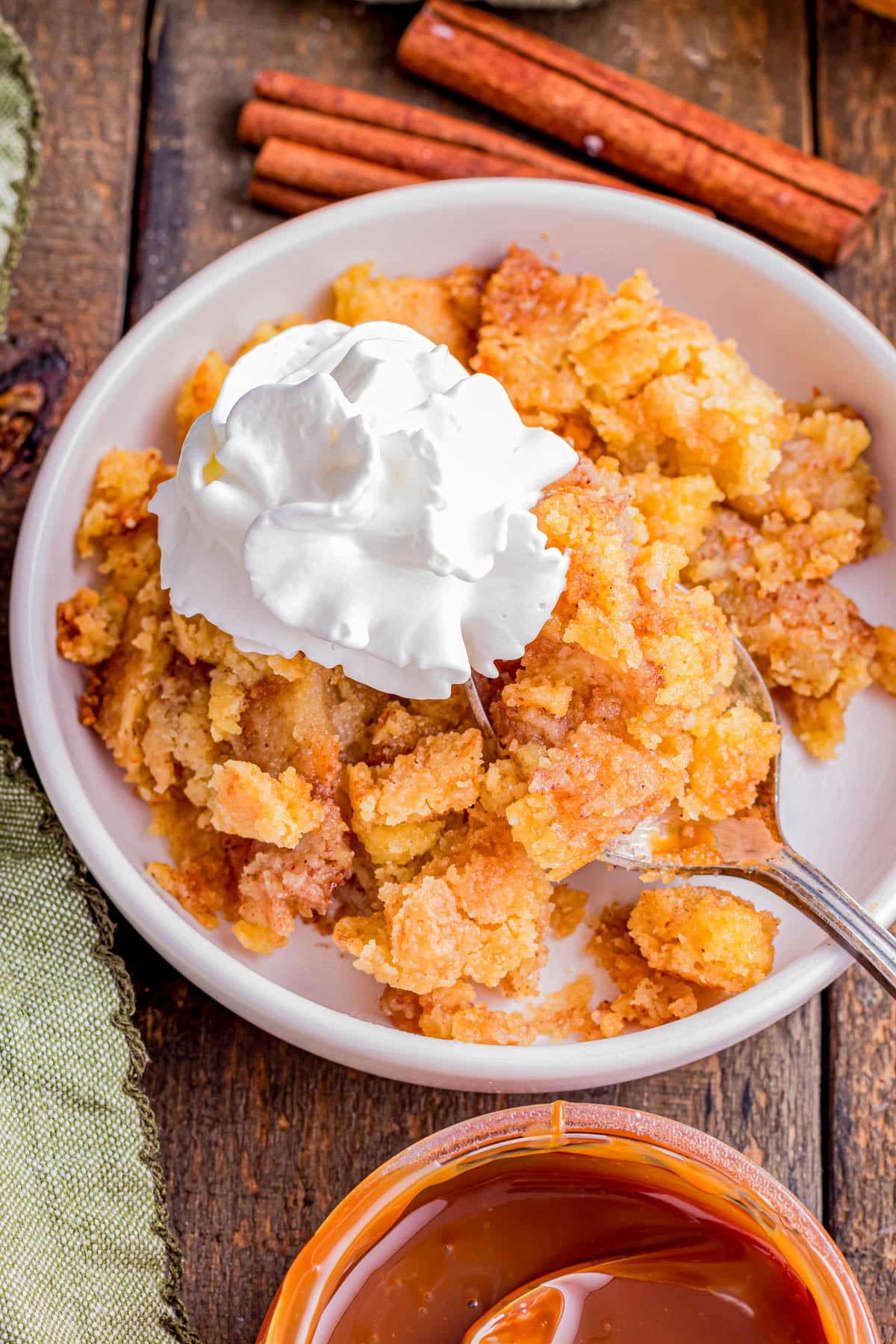 A plate of apple cobbler topped with whipped cream, with a fork, cinnamon sticks in the background, and a bowl of caramel sauce nearby.