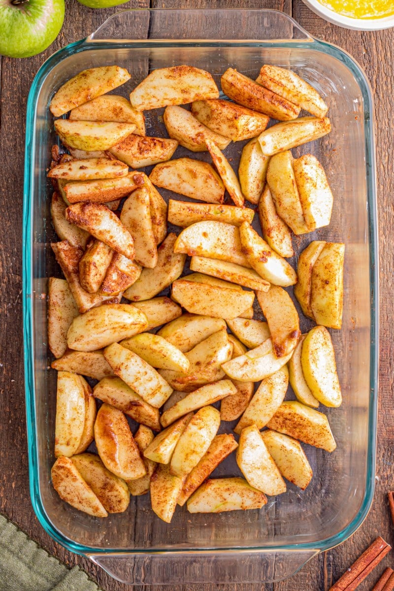 Sliced apples coated with cinnamon and sugar are arranged in a clear glass baking dish on a wooden surface.