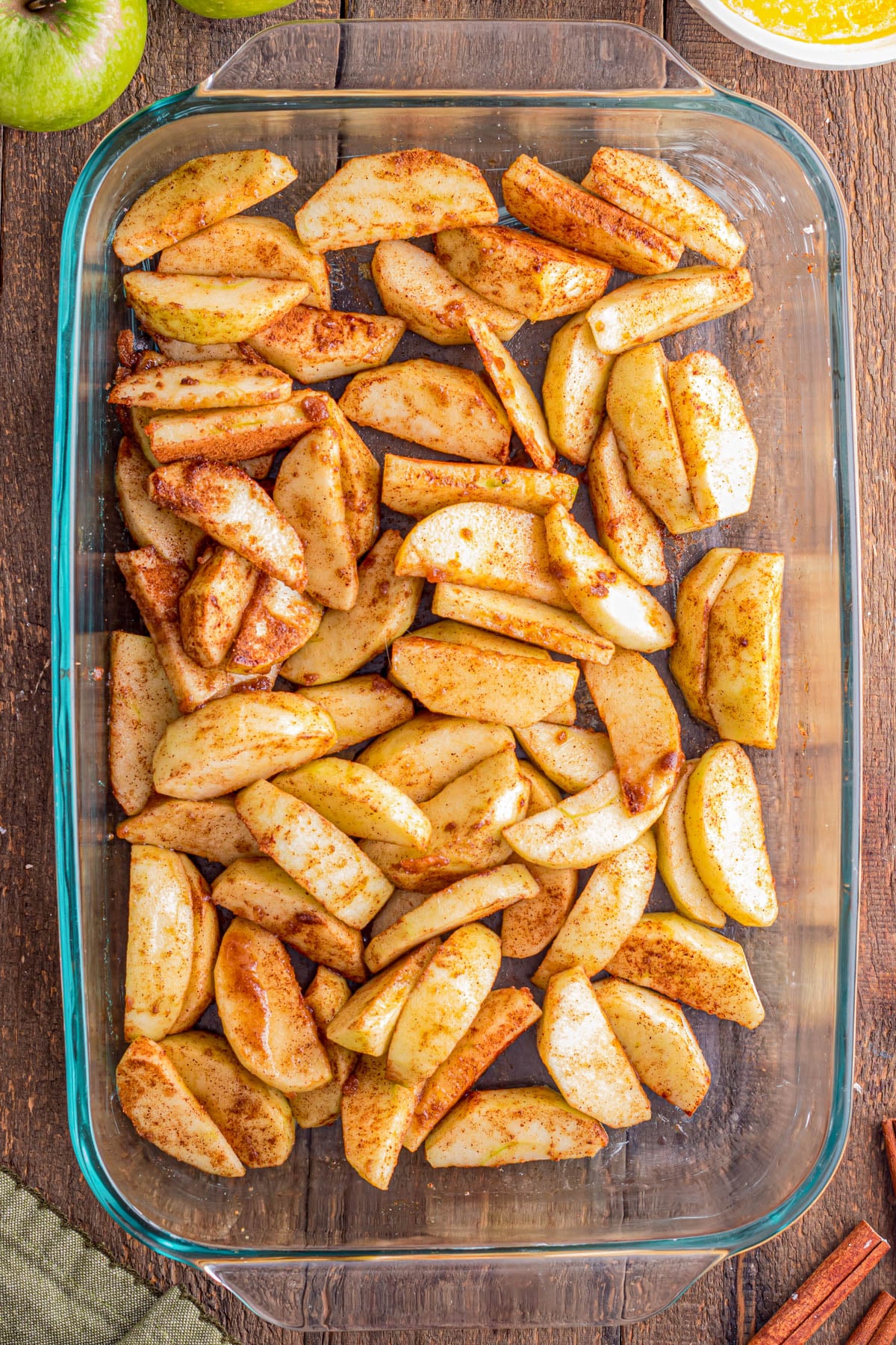 Sliced apples coated with cinnamon and sugar are arranged in a clear glass baking dish on a wooden surface.