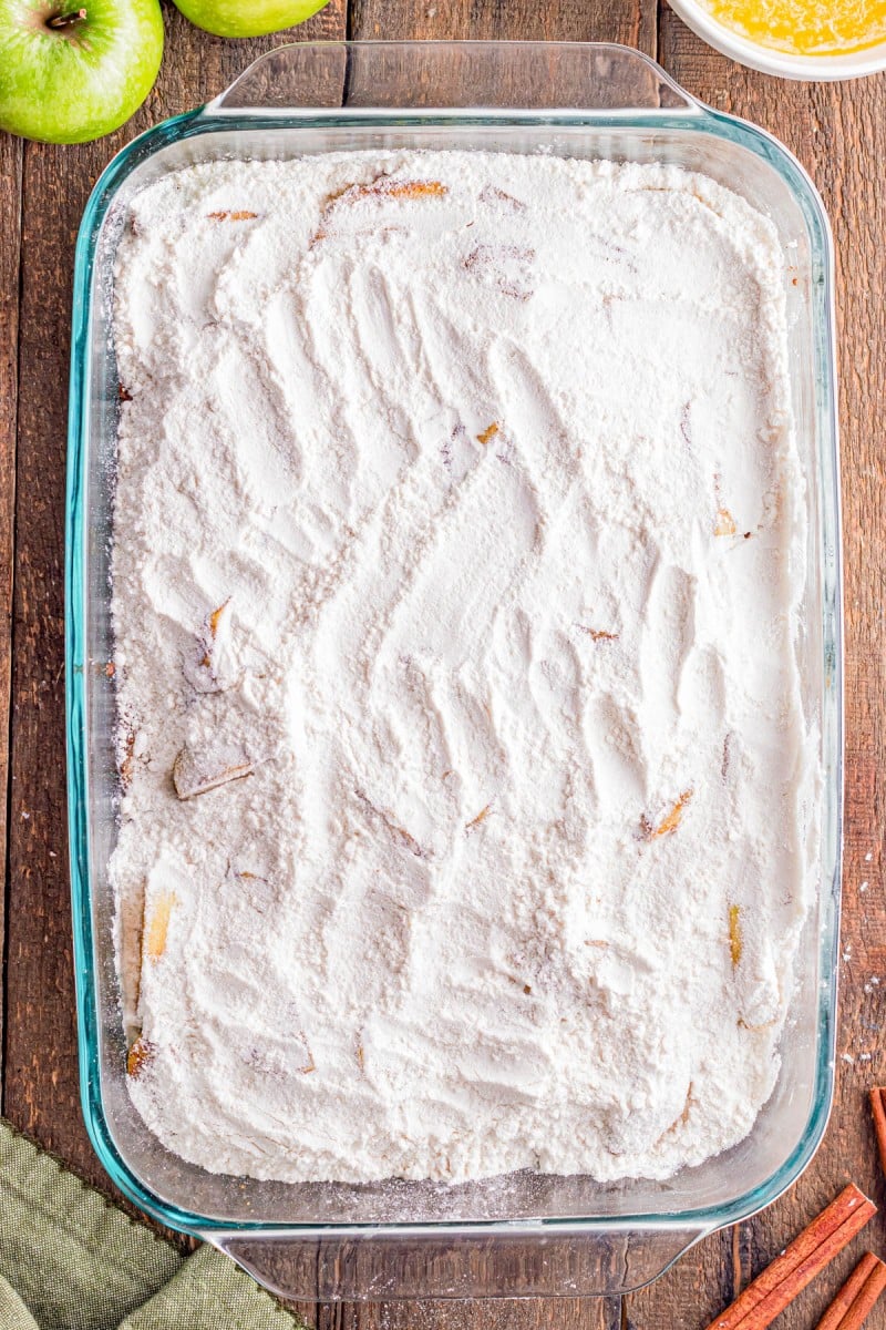 A glass baking dish filled with a layer of white flour mixture, showing visible finger marks and some apple slices partially covered.
