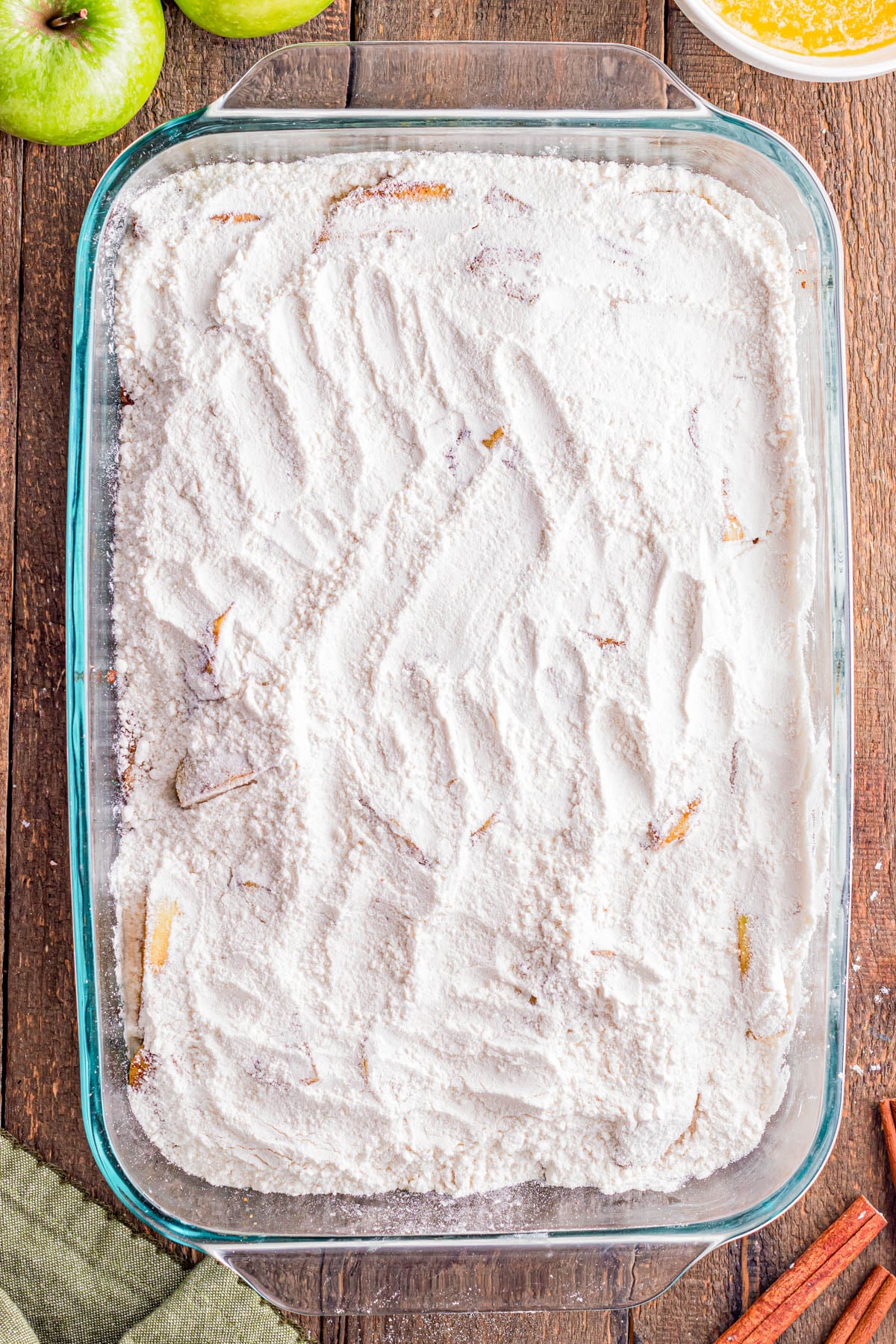 A glass baking dish filled with a layer of white flour mixture, showing visible finger marks and some apple slices partially covered.