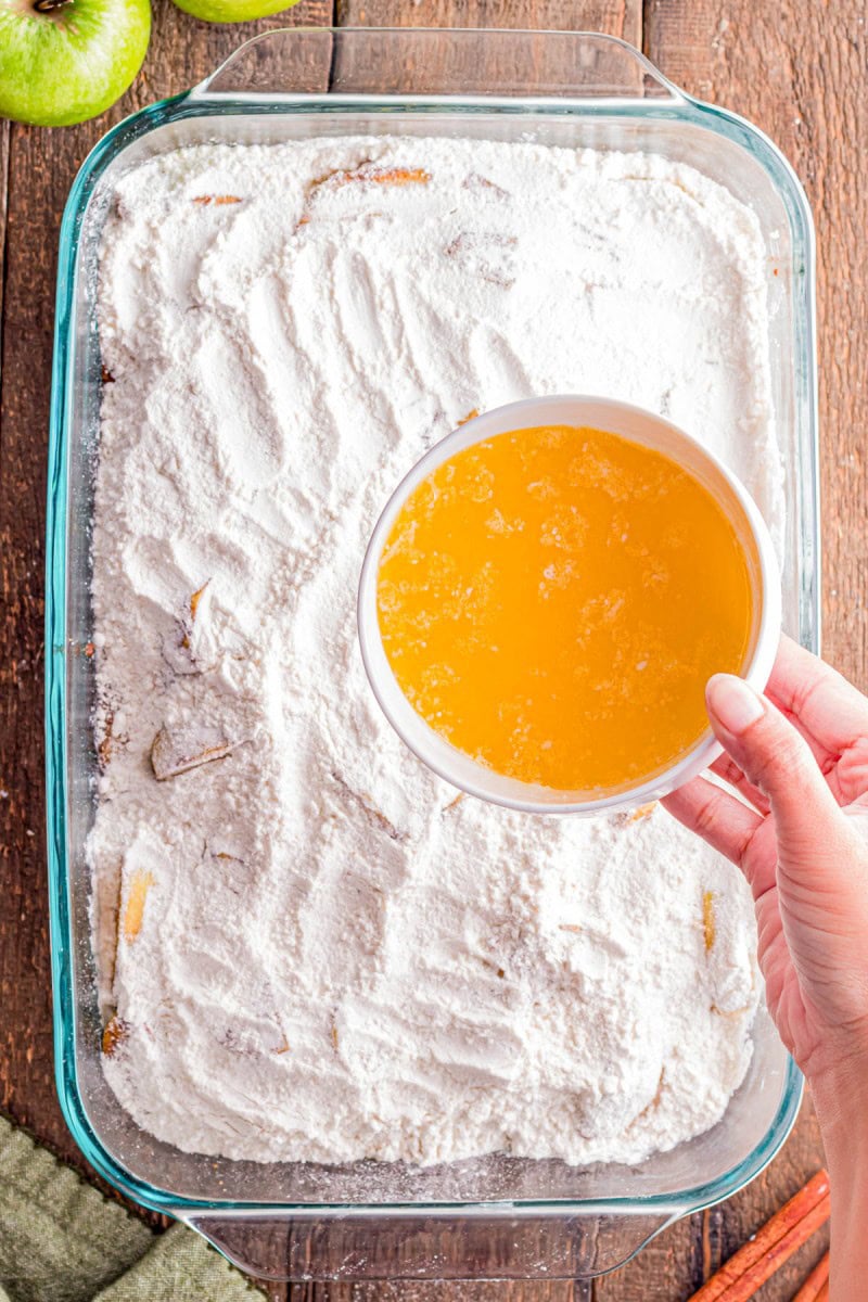 A hand pours melted butter from a white bowl over a baking dish filled with dry cake mix and apple slices on a wooden surface.