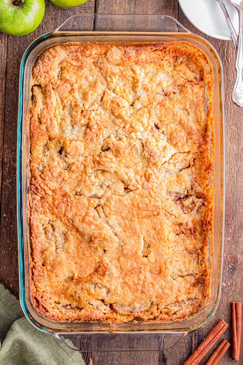 A baked apple cobbler in a rectangular glass dish on a wooden table, with green apples, cinnamon sticks, plates, and utensils nearby.