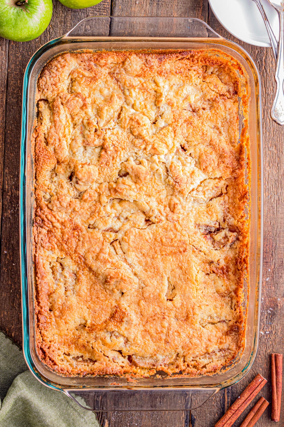 A baked apple cobbler in a rectangular glass dish on a wooden table, with green apples, cinnamon sticks, plates, and utensils nearby.