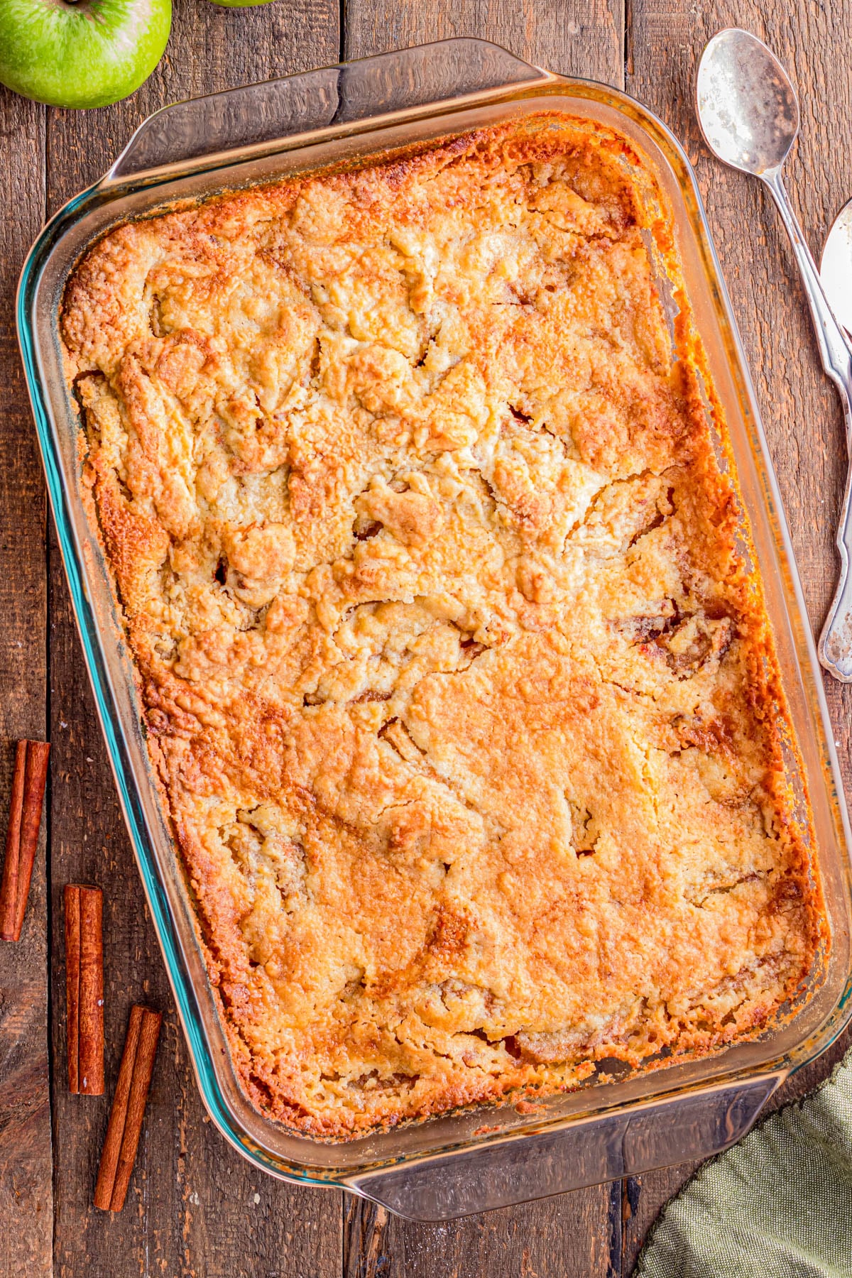 Glass baking dish filled with golden-brown apple cobbler on a wooden table, with cinnamon sticks, an apple, and two spoons nearby.