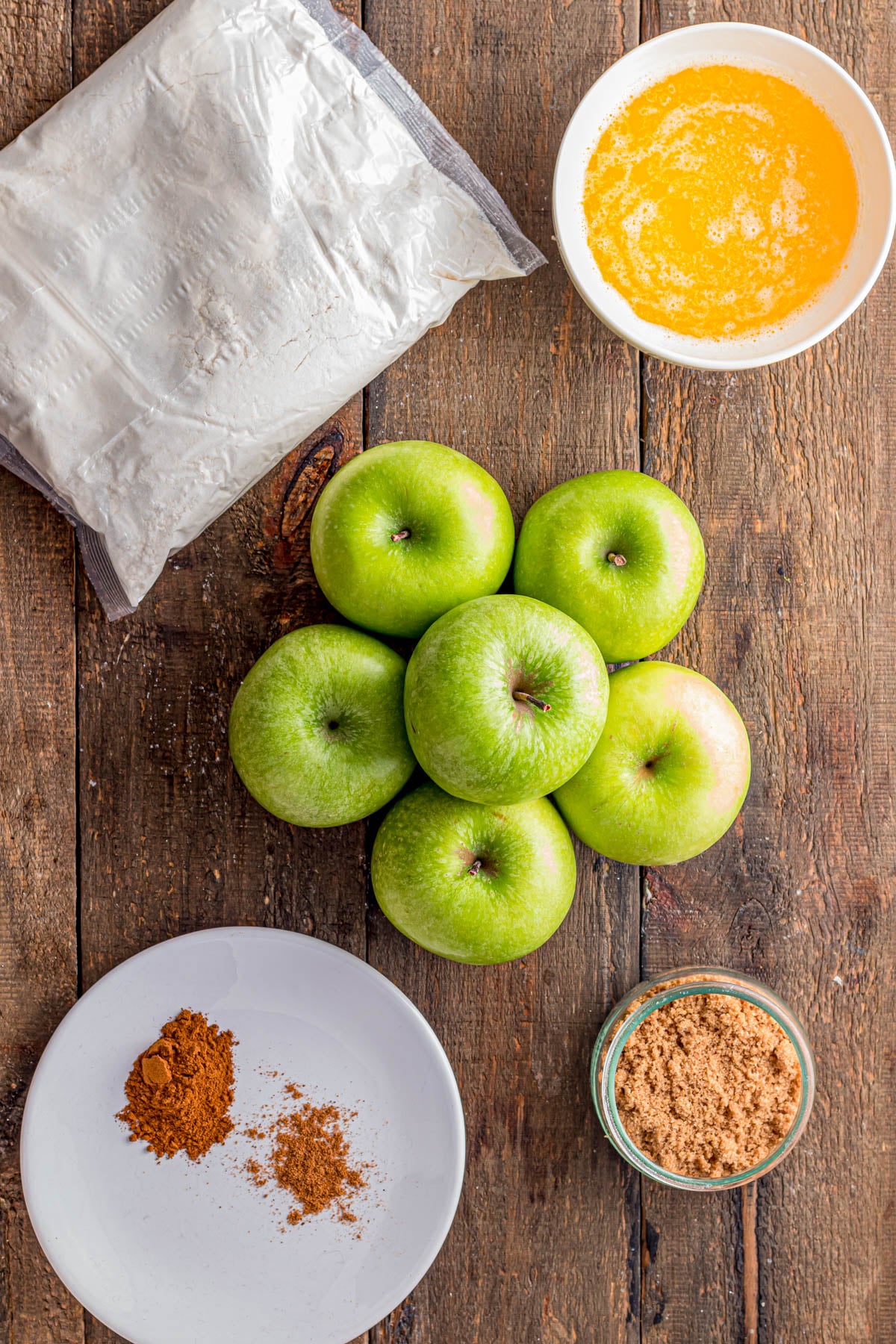 A bundle of green apples, cake mix, melted butter, brown sugar, and spices arranged on a wooden table.