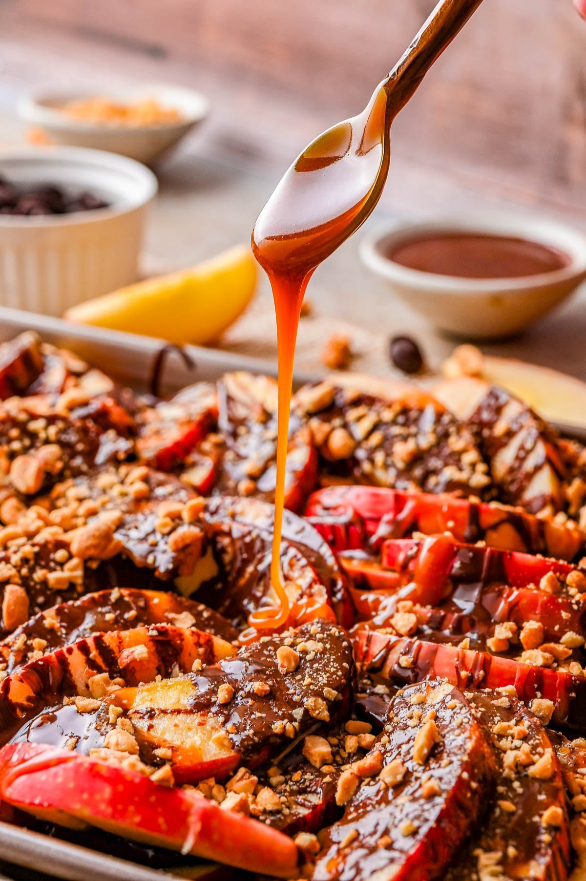 A spoon drizzles caramel sauce over sliced apples topped with chocolate and crushed nuts, with bowls of ingredients in the background.