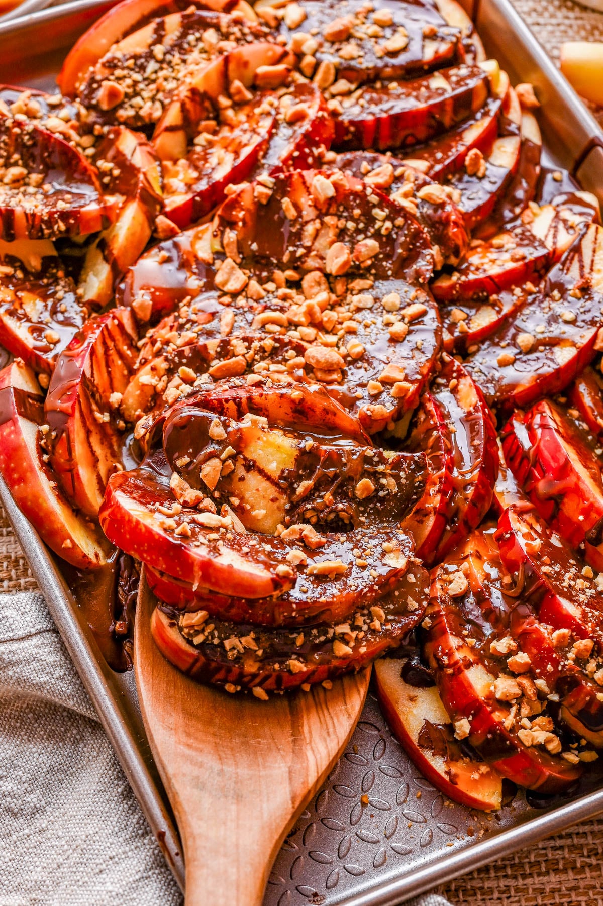 Sliced red apples arranged on a baking sheet, topped with chocolate drizzle and chopped nuts, with a wooden spatula lifting some slices.