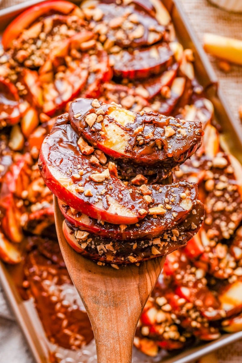 Sliced apples coated in chocolate and sprinkled with chopped nuts are lifted by a wooden spoon, with more arranged on a tray in the background.
