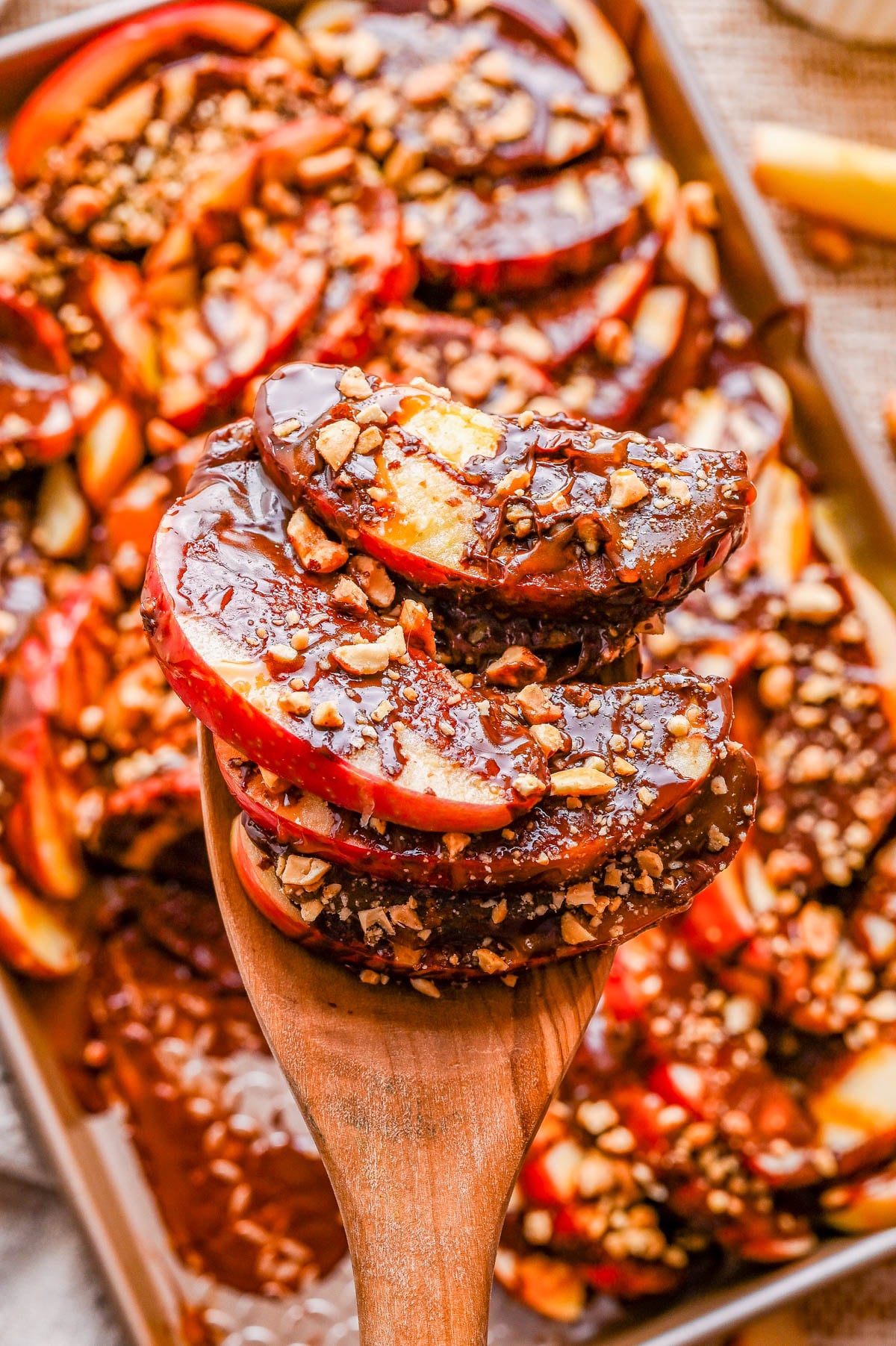 Sliced apples coated in chocolate and sprinkled with chopped nuts are lifted by a wooden spoon, with more arranged on a tray in the background.