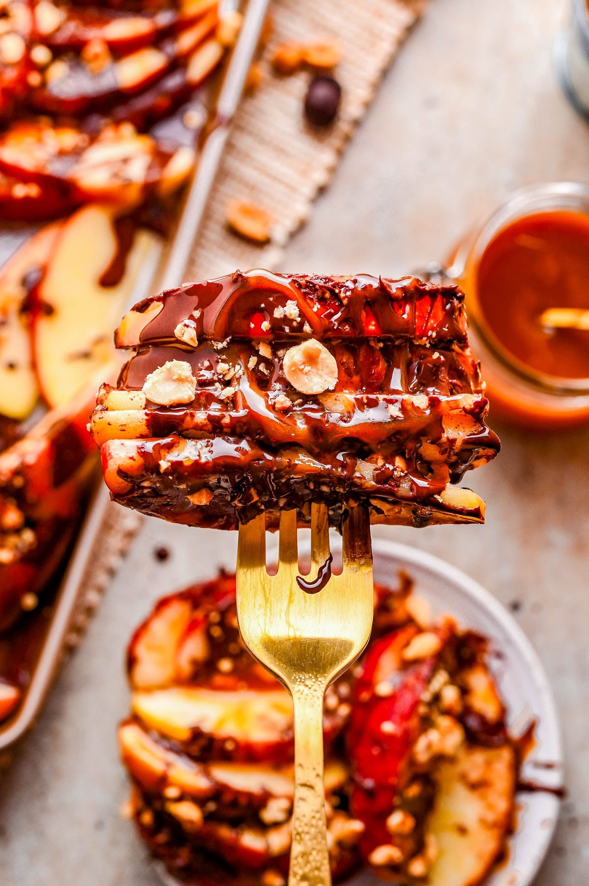 A close-up of a fork holding sliced apples drizzled with chocolate and caramel, topped with chopped nuts, with more apple slices and sauces in the background.