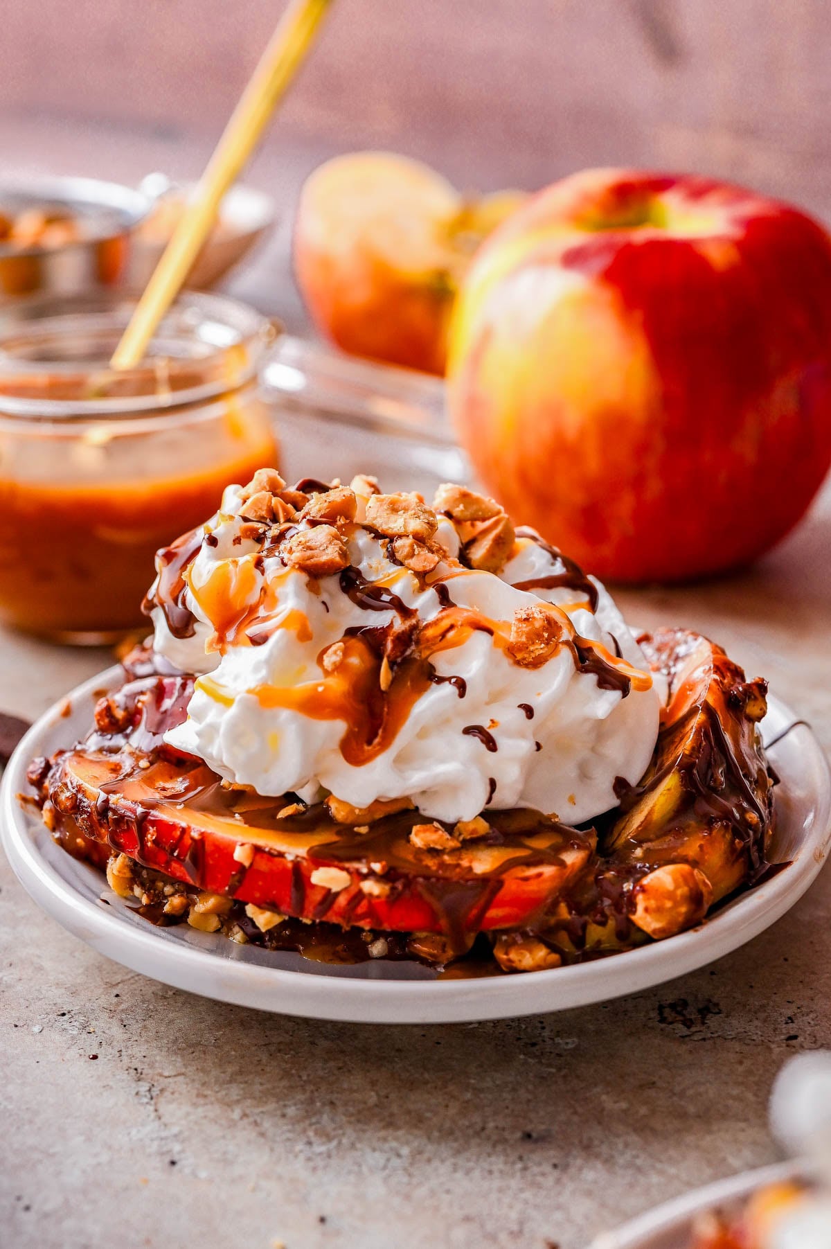 Apple slices topped with whipped cream, granola, chocolate, and caramel sauce are served on a small plate, with apples and caramel sauce jars in the background.