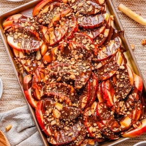 Sliced apples on a tray topped with chocolate drizzle and chopped nuts, with whole and sliced apples visible nearby.