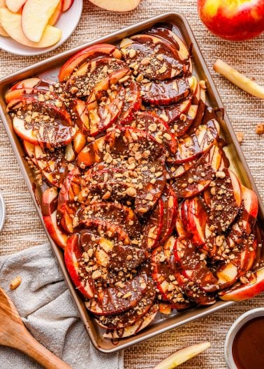 Sliced apples on a tray topped with chocolate drizzle and chopped nuts, with whole and sliced apples visible nearby.