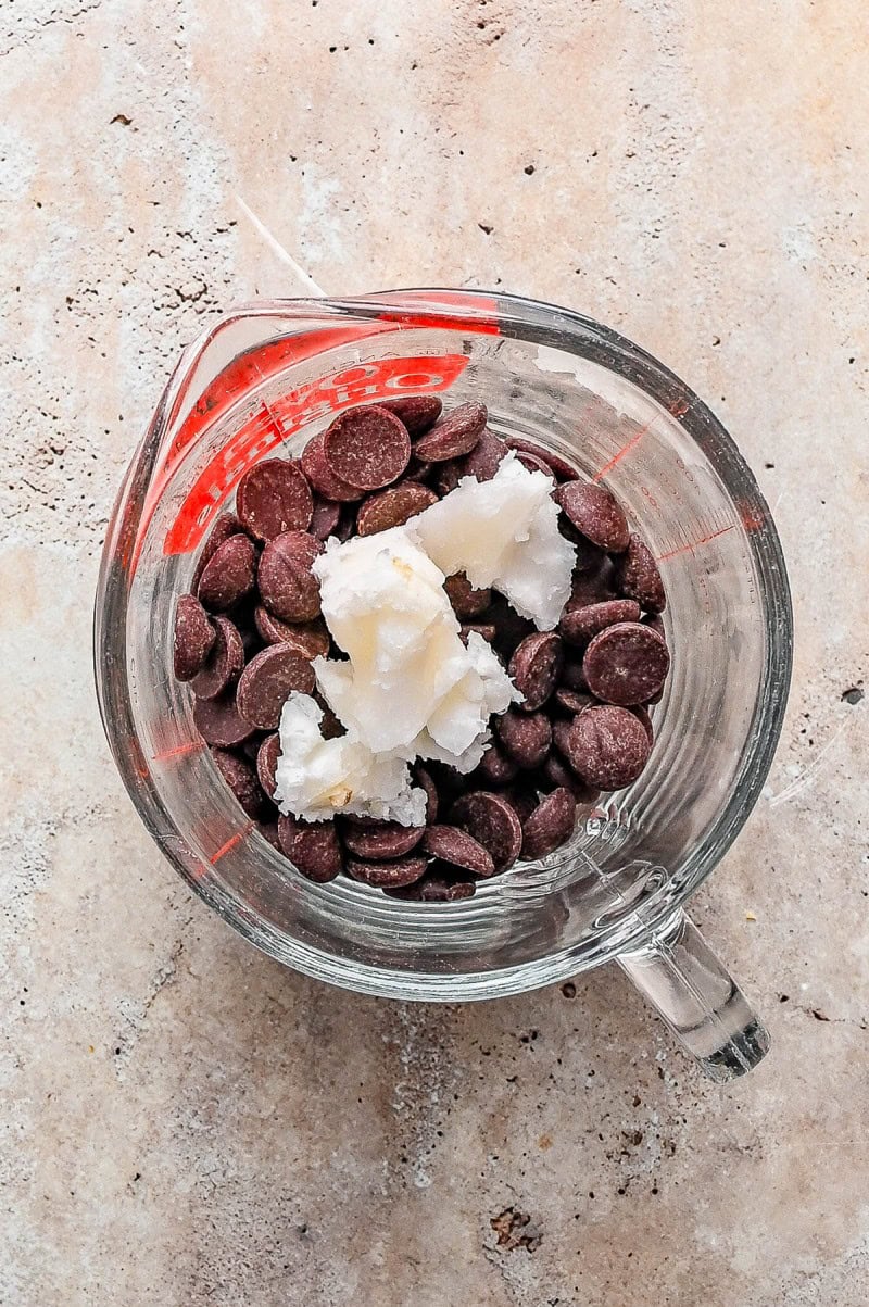 A glass measuring cup filled with chocolate chips and dollops of solid coconut oil on a light-colored surface.