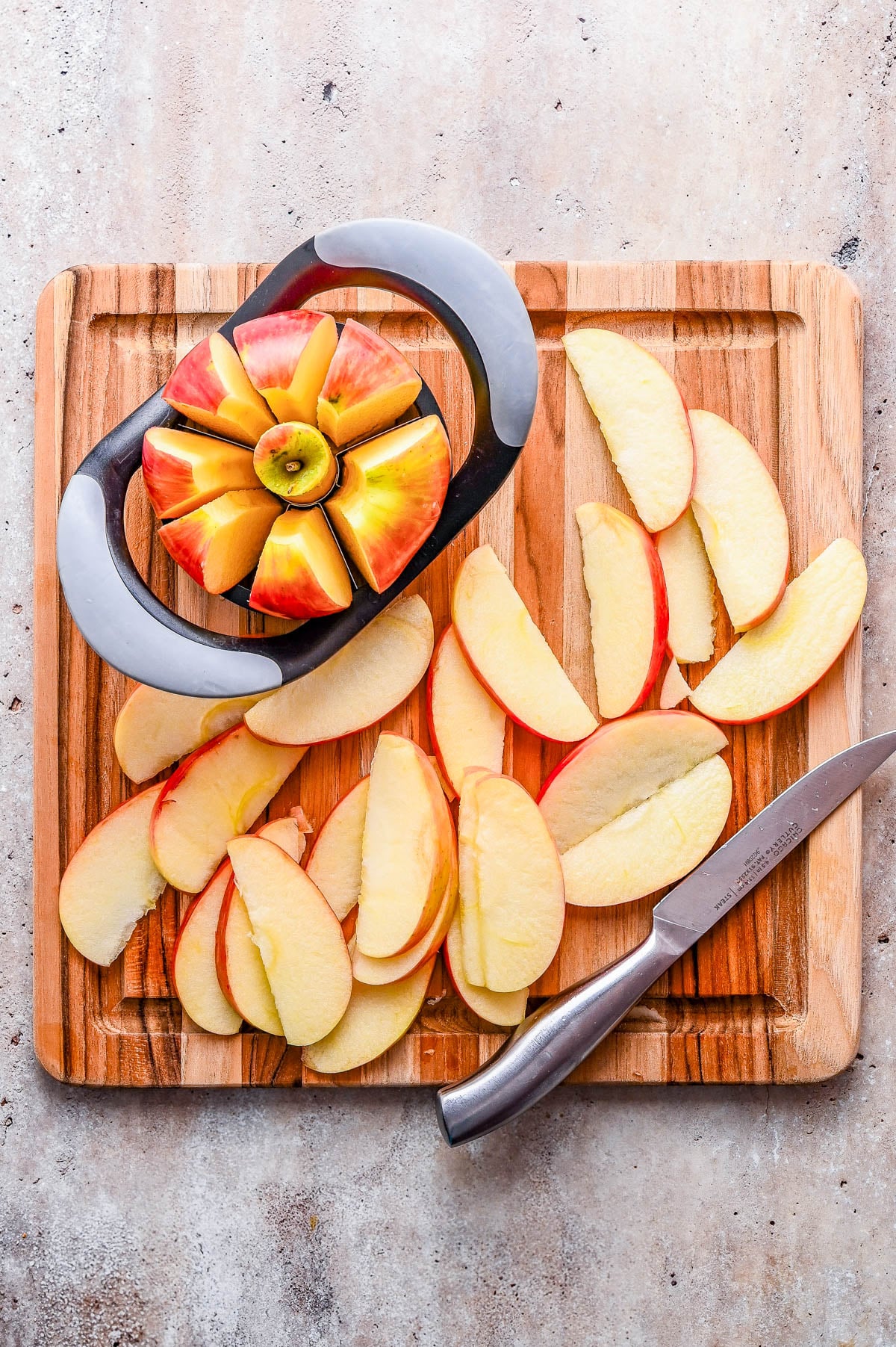 Apple slices on a wooden cutting board with an apple slicer and a knife; some apple pieces remain in the slicer.