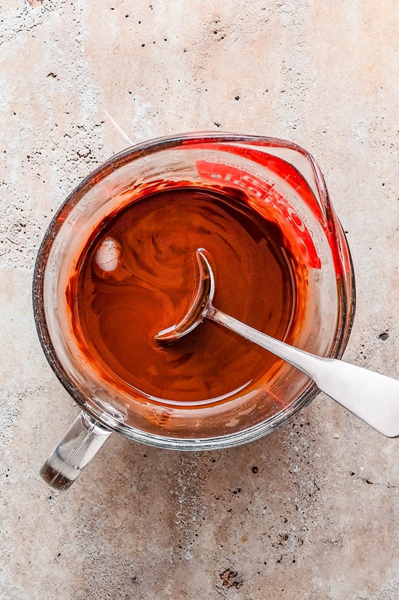 Glass measuring cup filled with melted chocolate and a metal spoon resting inside, placed on a light textured surface.