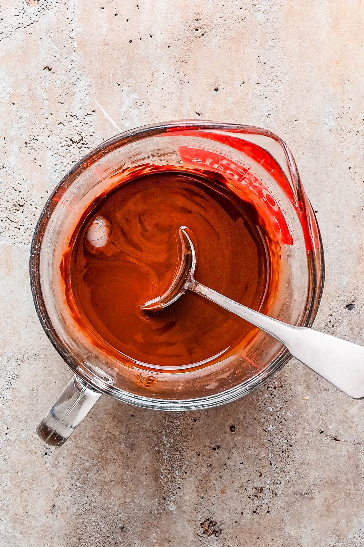 Glass measuring cup filled with melted chocolate and a metal spoon resting inside, placed on a light textured surface.