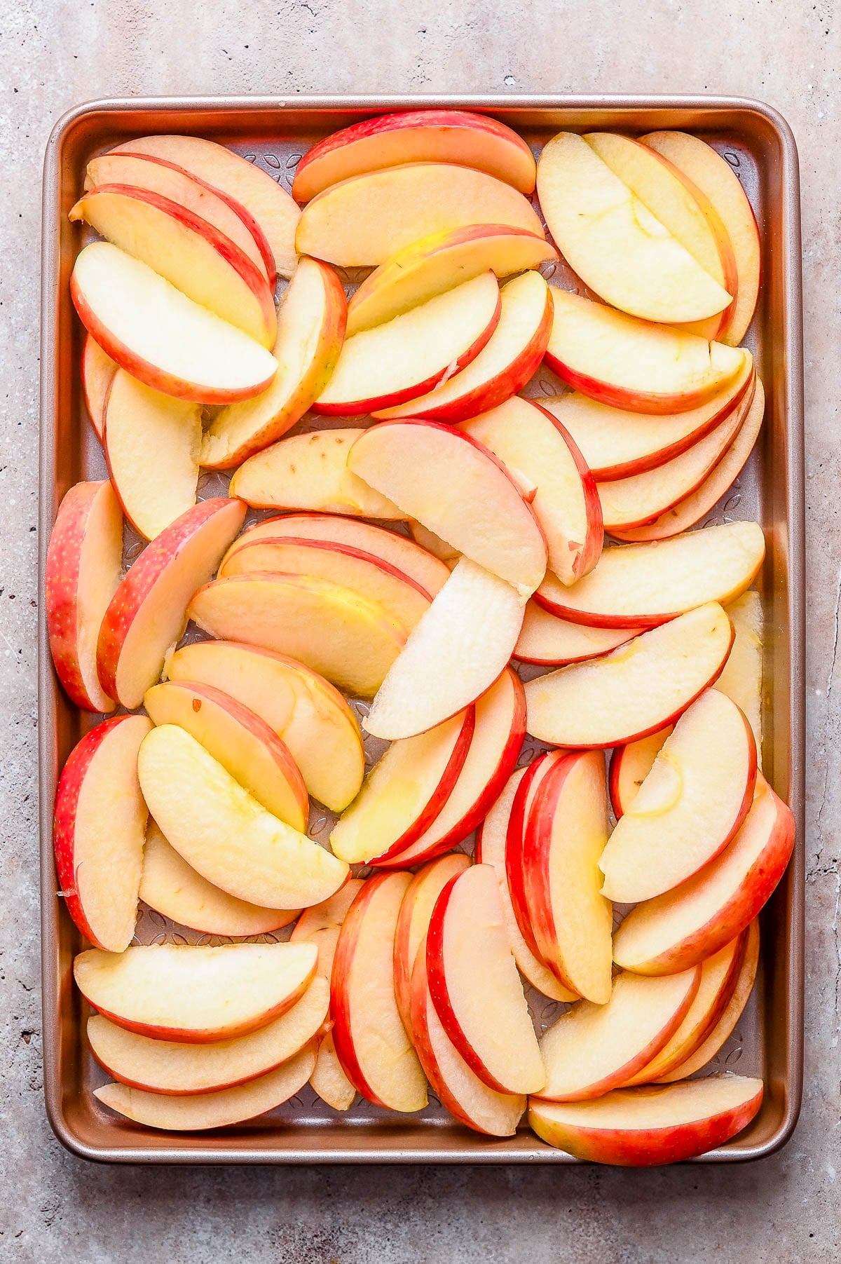 Baking sheet with evenly spread raw apple slices, arranged in a single layer on a light, textured surface.