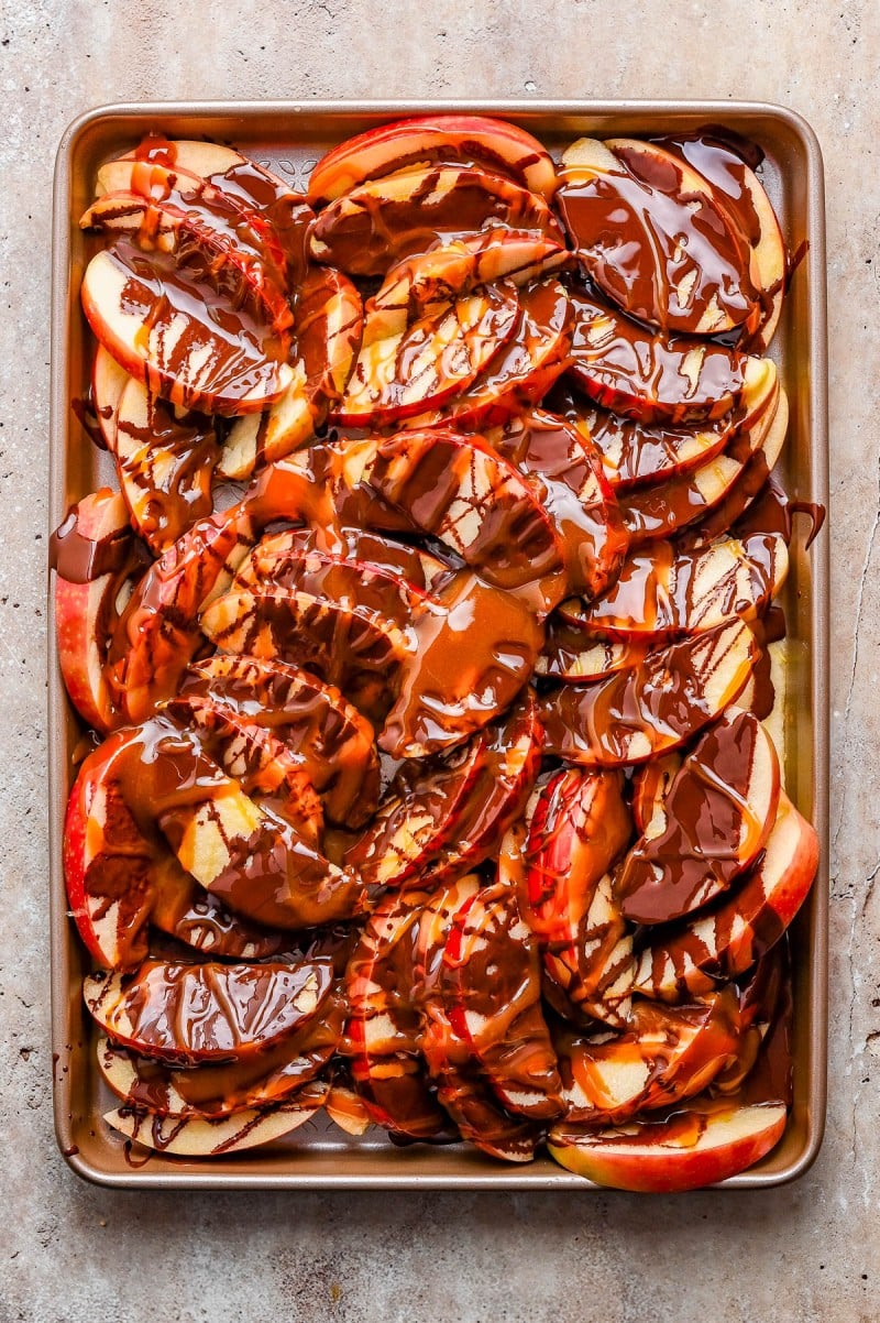 Baking sheet filled with sliced apples drizzled generously with melted chocolate on a neutral-colored surface.