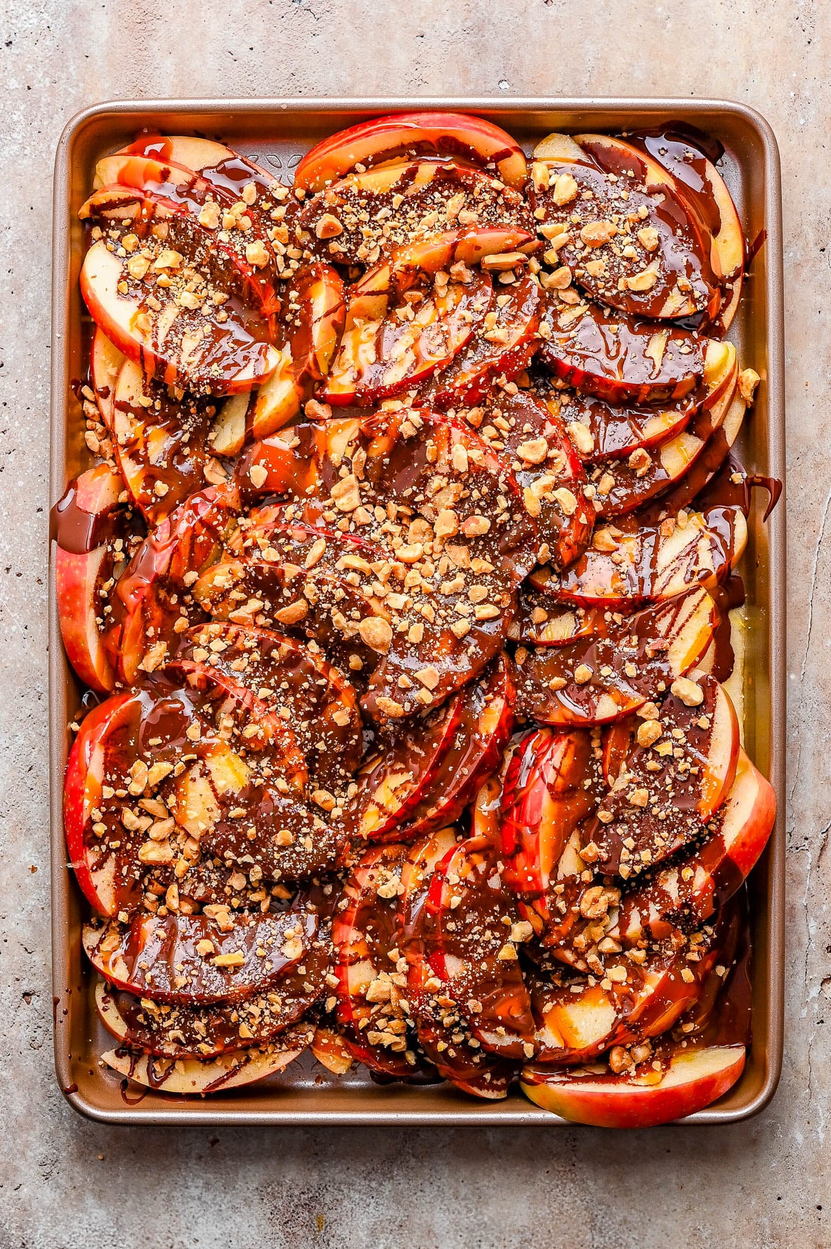Sliced apples on a baking sheet topped with drizzled chocolate and crushed nuts.