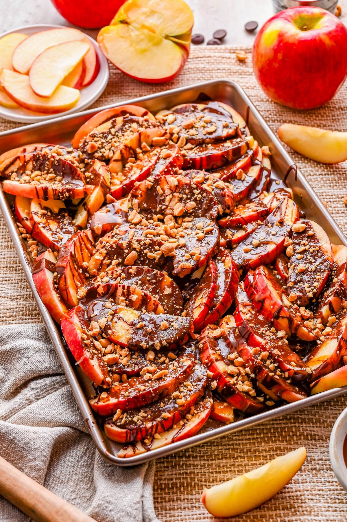 A baking sheet filled with sliced apples topped with chocolate drizzle, chopped nuts, and toffee bits; whole and sliced apples are arranged nearby.