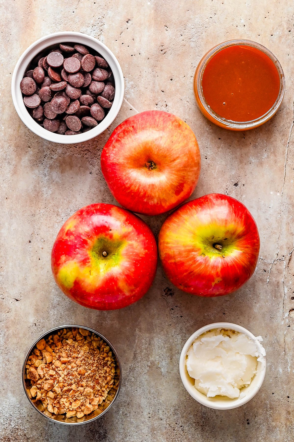 Three apples, a bowl of chocolate chips, a jar of caramel sauce, chopped nuts, and a bowl of white cream on a beige surface.