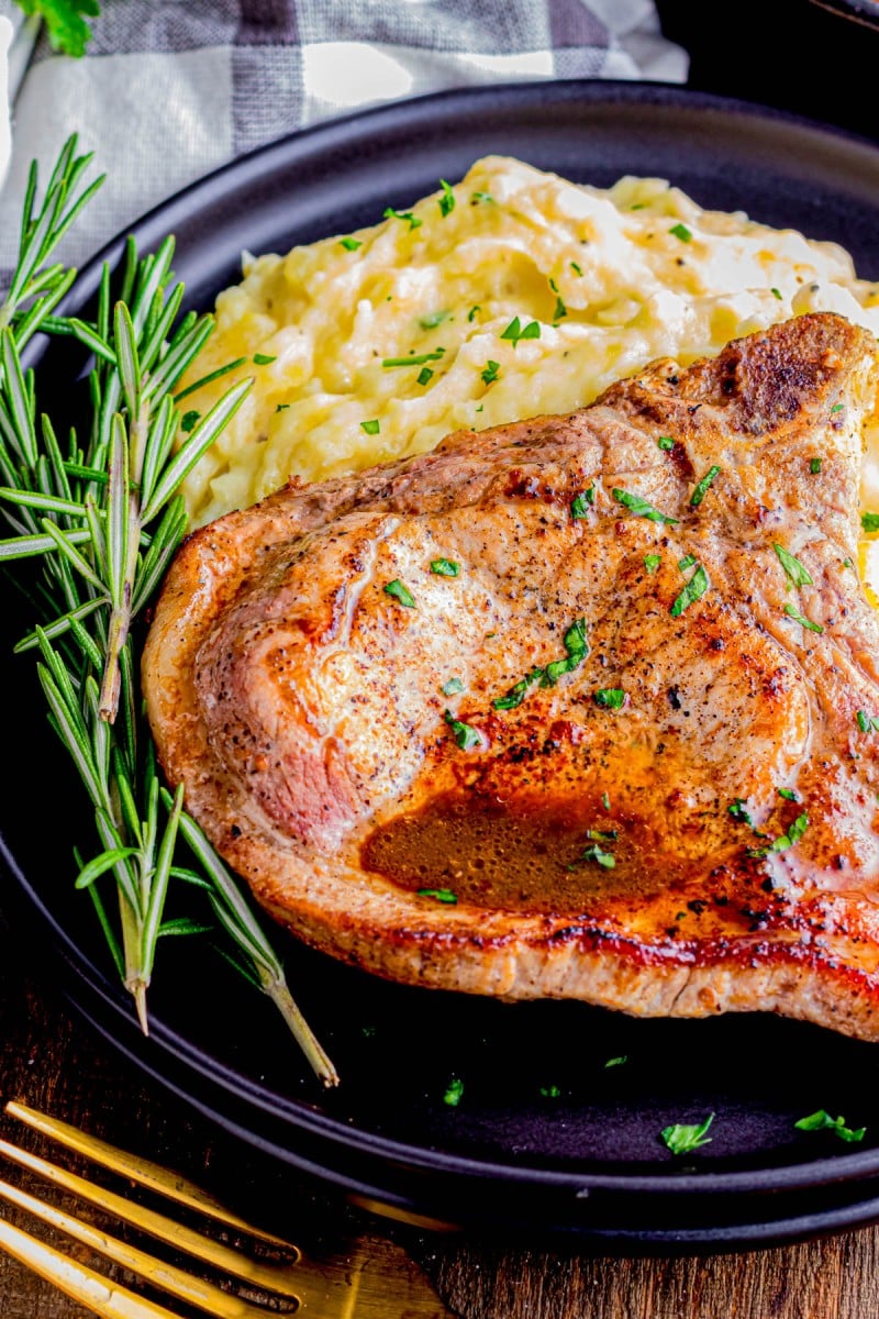 A cooked pork chop garnished with herbs sits beside creamy mashed potatoes on a black plate, with sprigs of fresh rosemary on the side.
