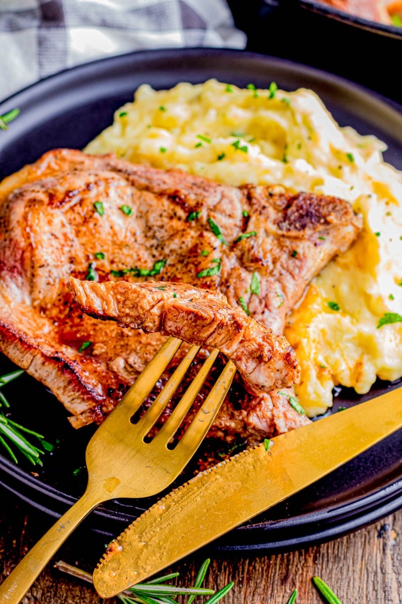 A cooked steak with a slice cut sits on a plate next to a serving of mashed potatoes. A gold fork and knife are placed on the edge of the plate.