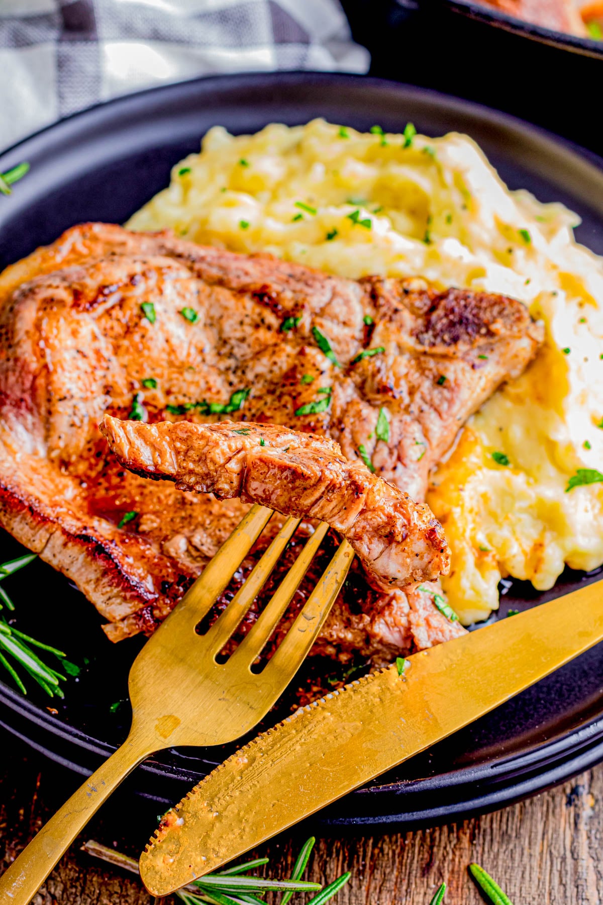 A cooked steak with a slice cut sits on a plate next to a serving of mashed potatoes. A gold fork and knife are placed on the edge of the plate.