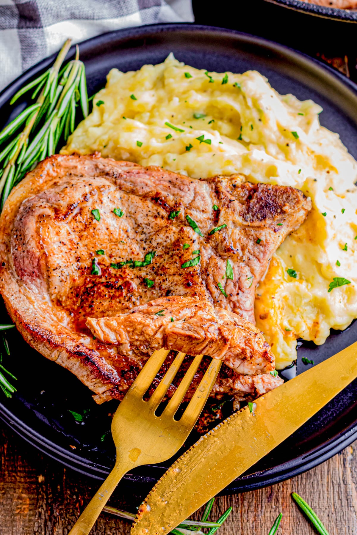 Grilled pork chop served with mashed potatoes on a black plate, garnished with rosemary. A gold fork and knife rest next to the food.