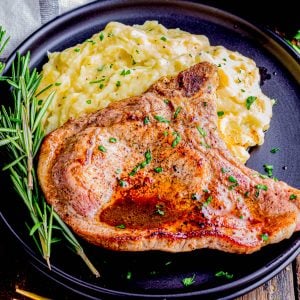 A cooked pork chop garnished with herbs is served with mashed potatoes on a black plate, next to a sprig of rosemary and a gold fork and knife.
