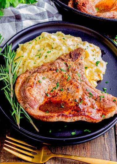 A cooked pork chop garnished with herbs is served with mashed potatoes on a black plate, next to a sprig of rosemary and a gold fork and knife.